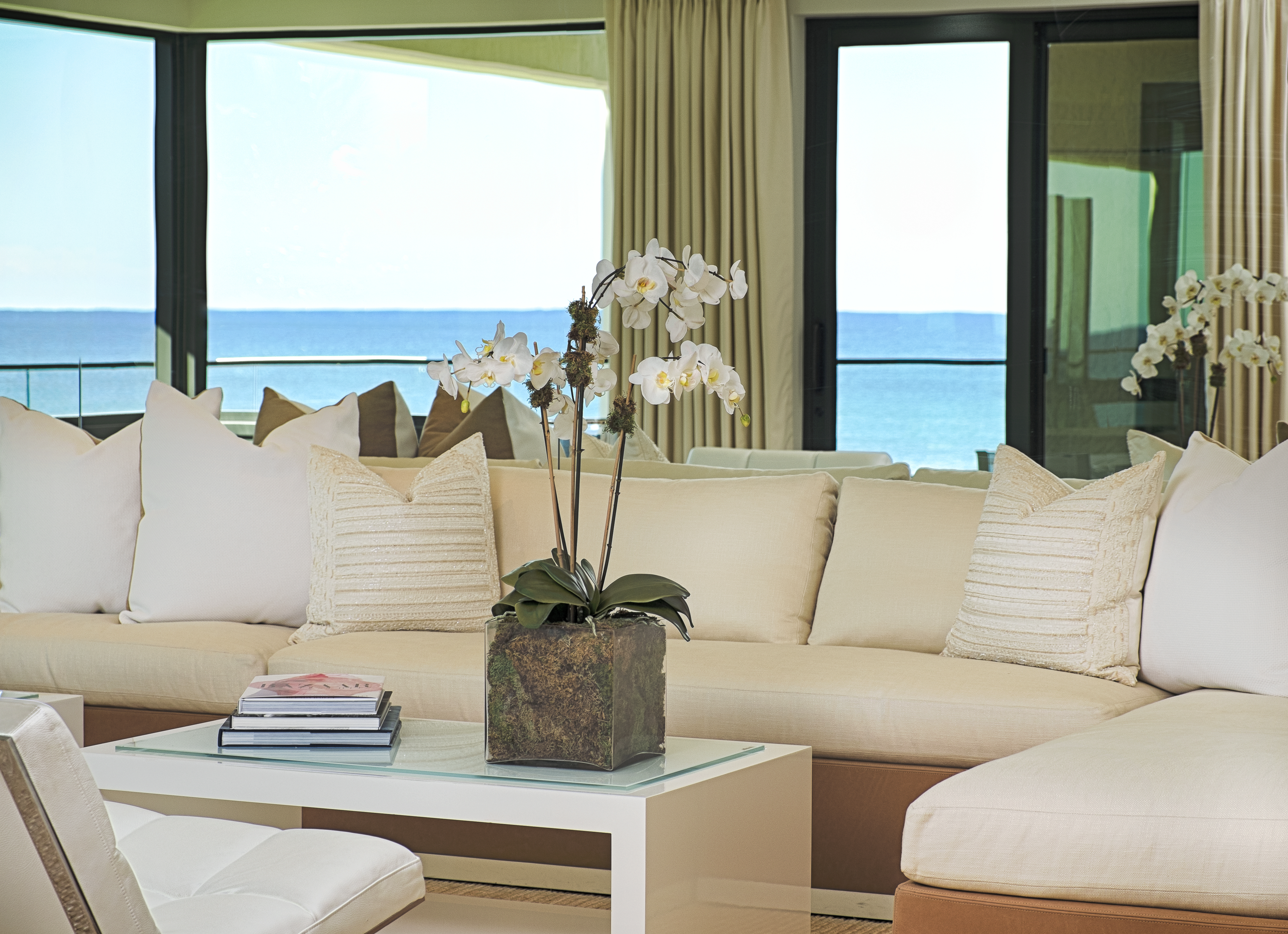Modern beachfront living room with beige sofas, white orchids, and ocean view through large windows.