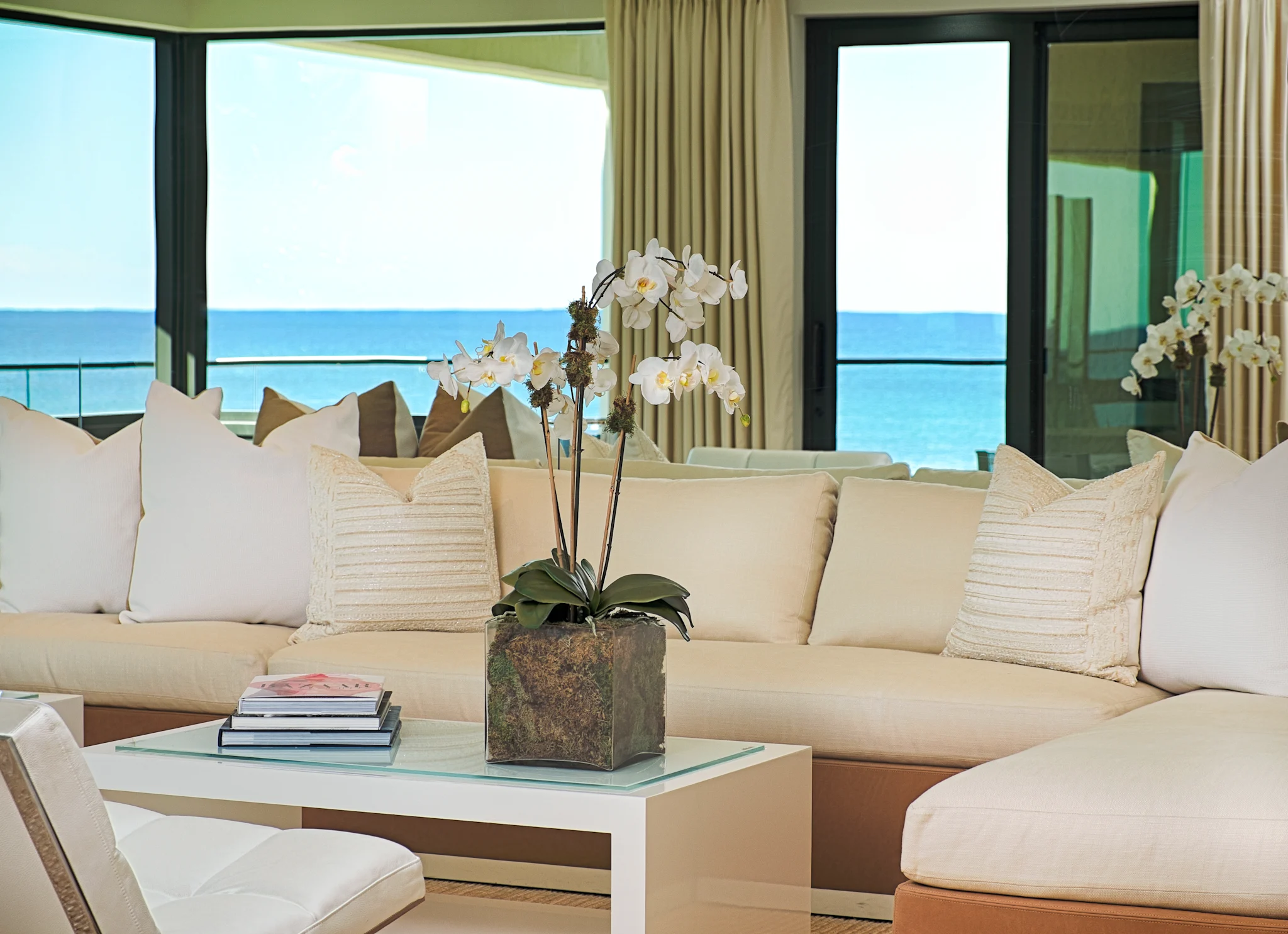 Modern beachfront living room with beige sofas, white orchids, and ocean view through large windows.