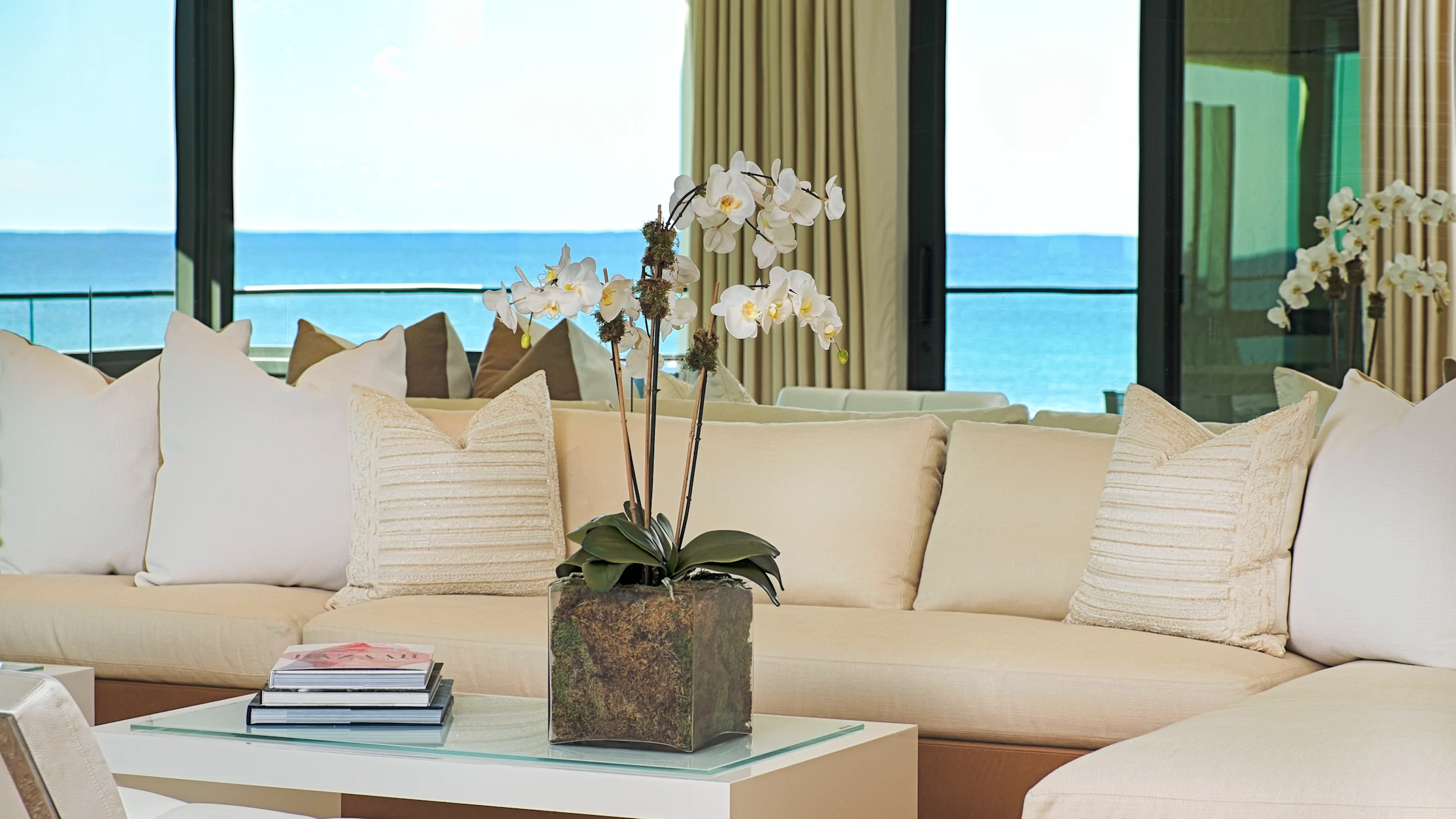 Modern beachfront living room with beige sofas, white orchids, and ocean view through large windows.