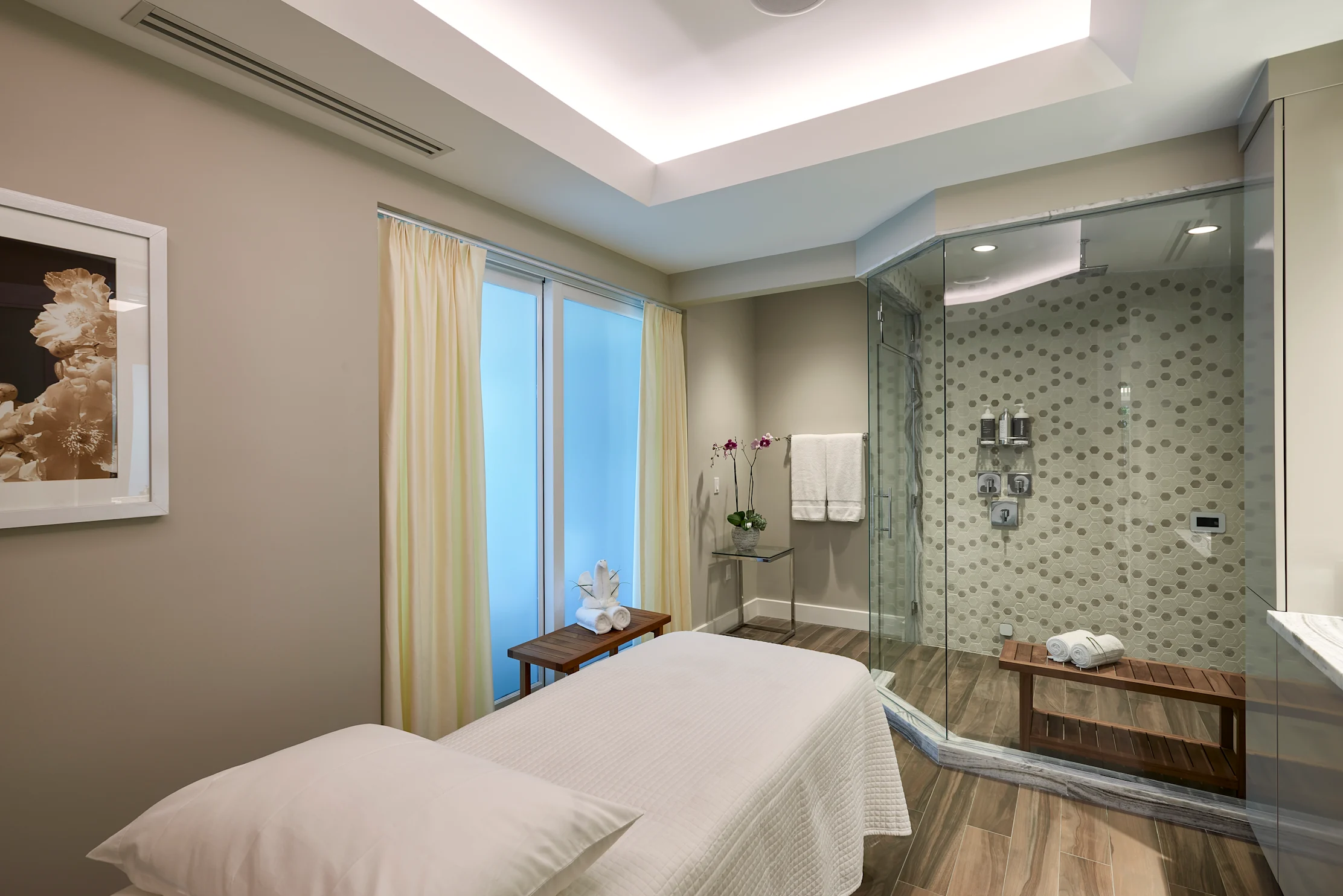 Modern spa treatment room with white massage table, glass-enclosed shower with mosaic tile, and ocean view.