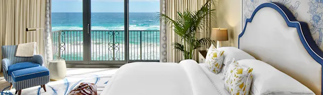 Elegant hotel bedroom with white bed, blue accents, and ocean view through balcony doors.