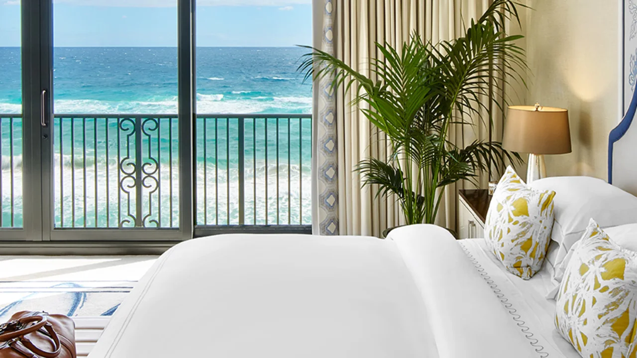 Elegant hotel bedroom with white bed, blue accents, and ocean view through balcony doors.