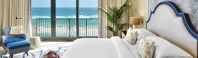 Elegant hotel bedroom with white bed, blue accents, and ocean view through balcony doors.