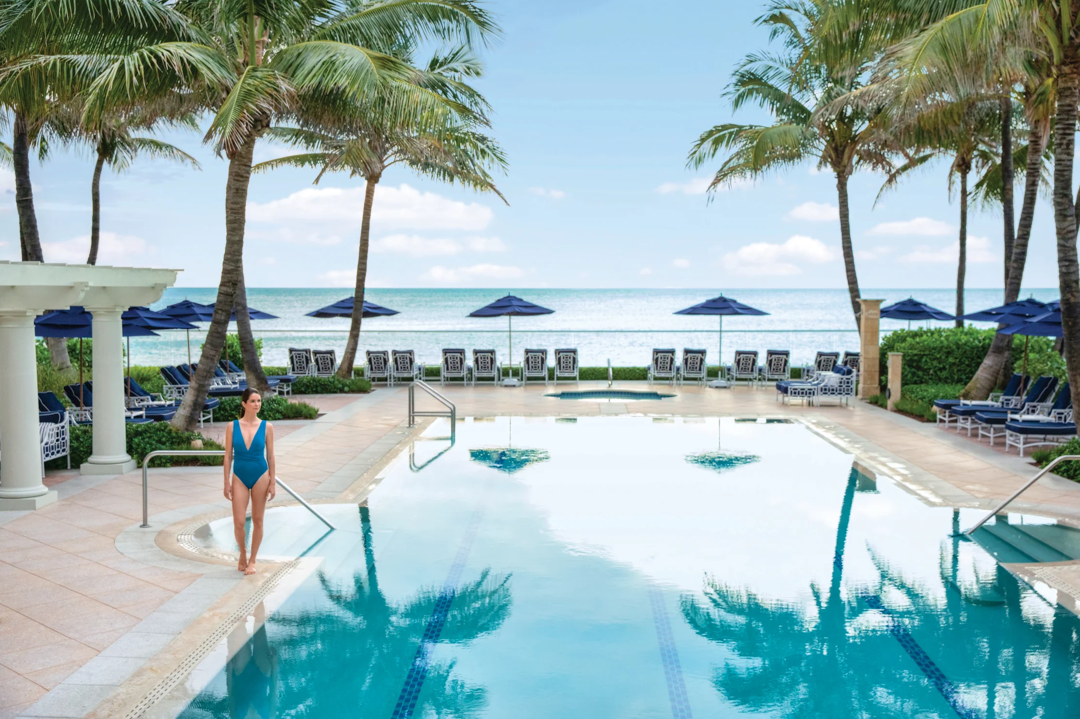Woman in blue swimsuit by luxury beachfront pool with palm trees and ocean view.