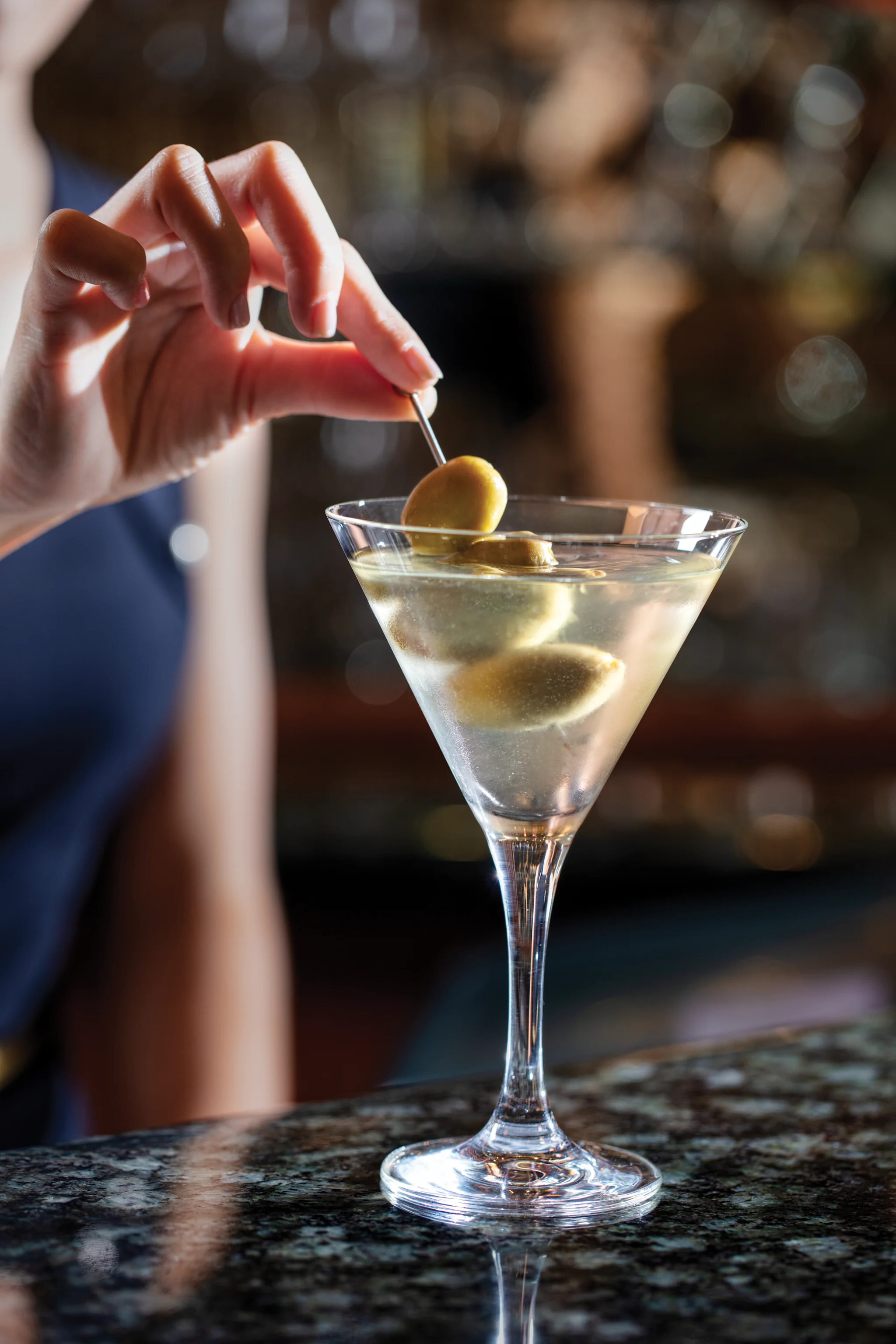Hand placing olive on toothpick into martini glass with clear cocktail on bar counter.