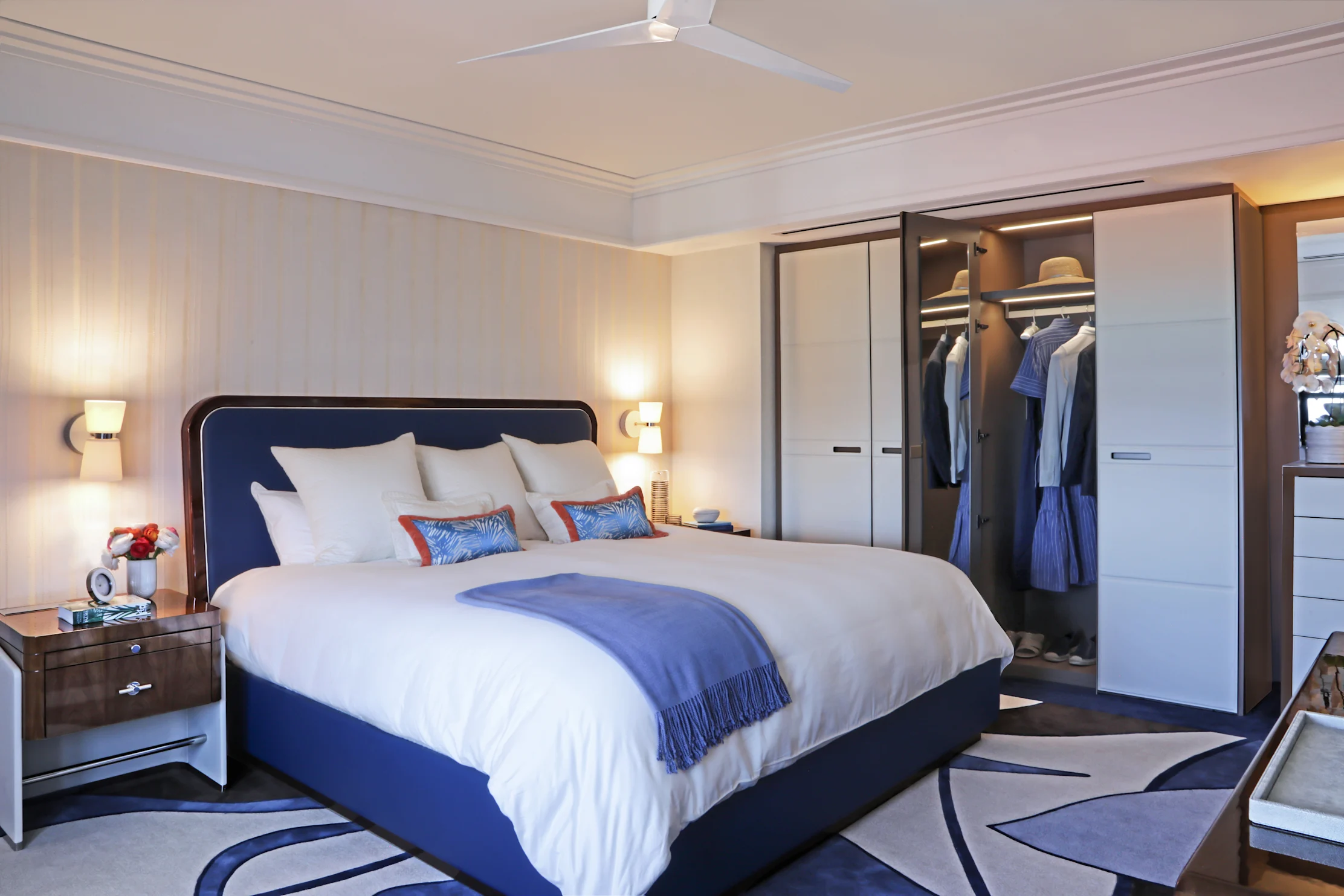 Modern hotel bedroom with navy blue upholstered bed, white linens, open wardrobe, and ceiling fan.