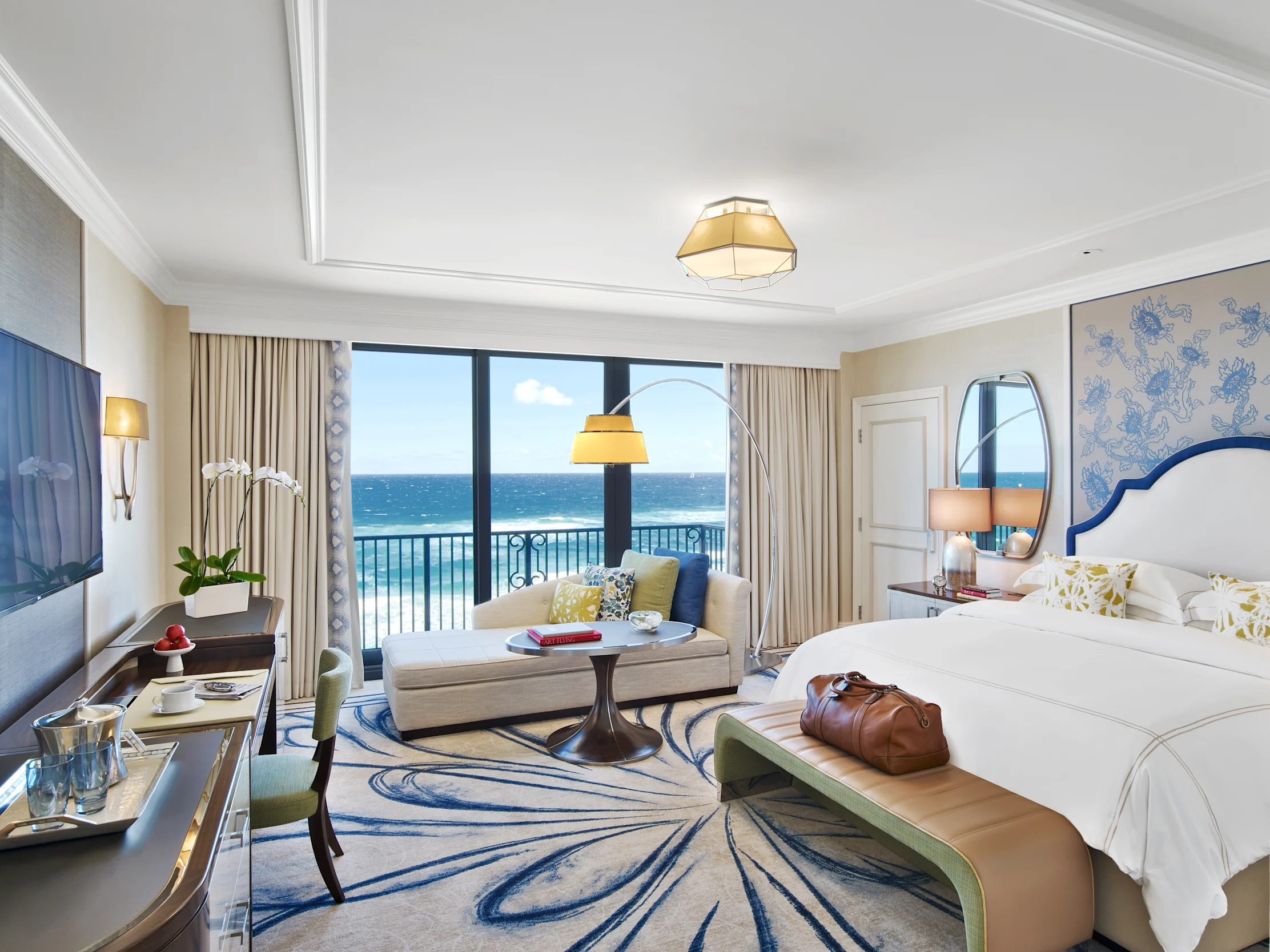 Elegant hotel room with ocean view, white bed, beige curtains, blue patterned rug, and modern decor.