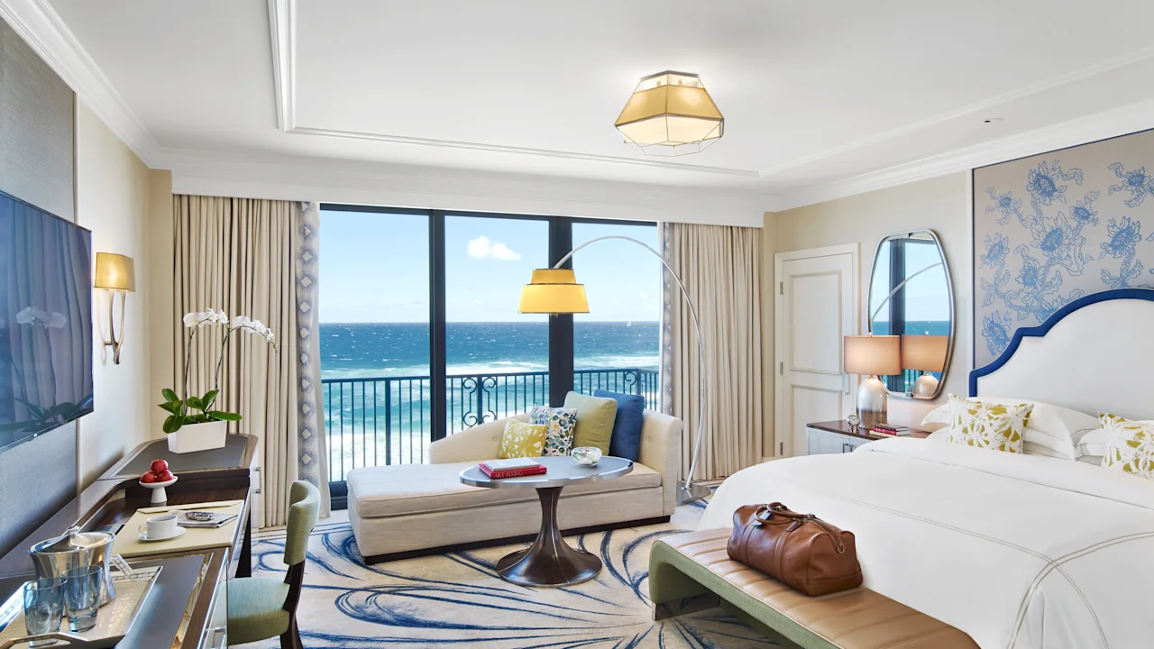 Elegant hotel room with ocean view, white bed, beige curtains, blue patterned rug, and modern decor.