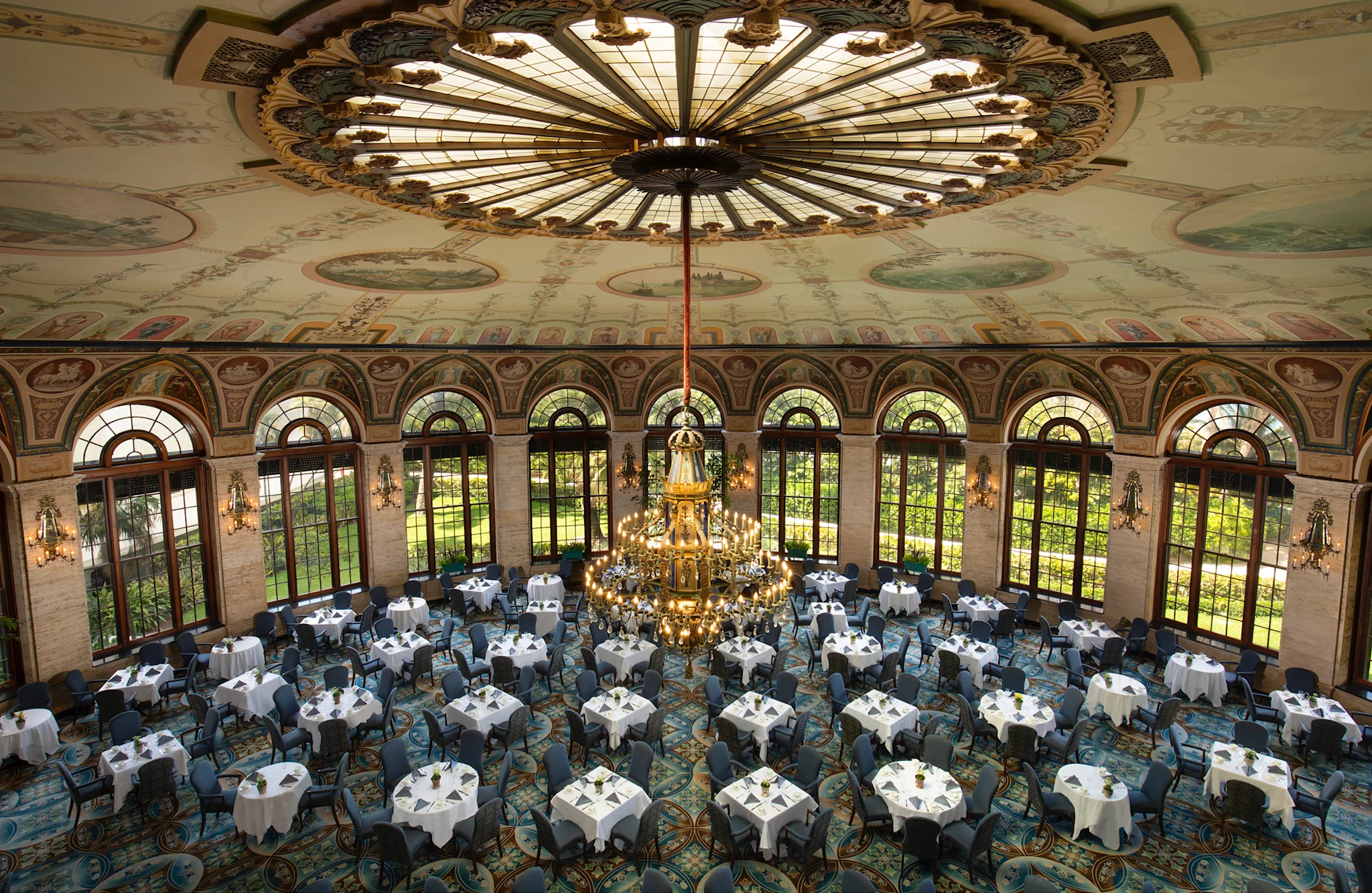 Ornate ballroom with decorative ceiling, large chandelier, arched windows, and round tables set for dining.