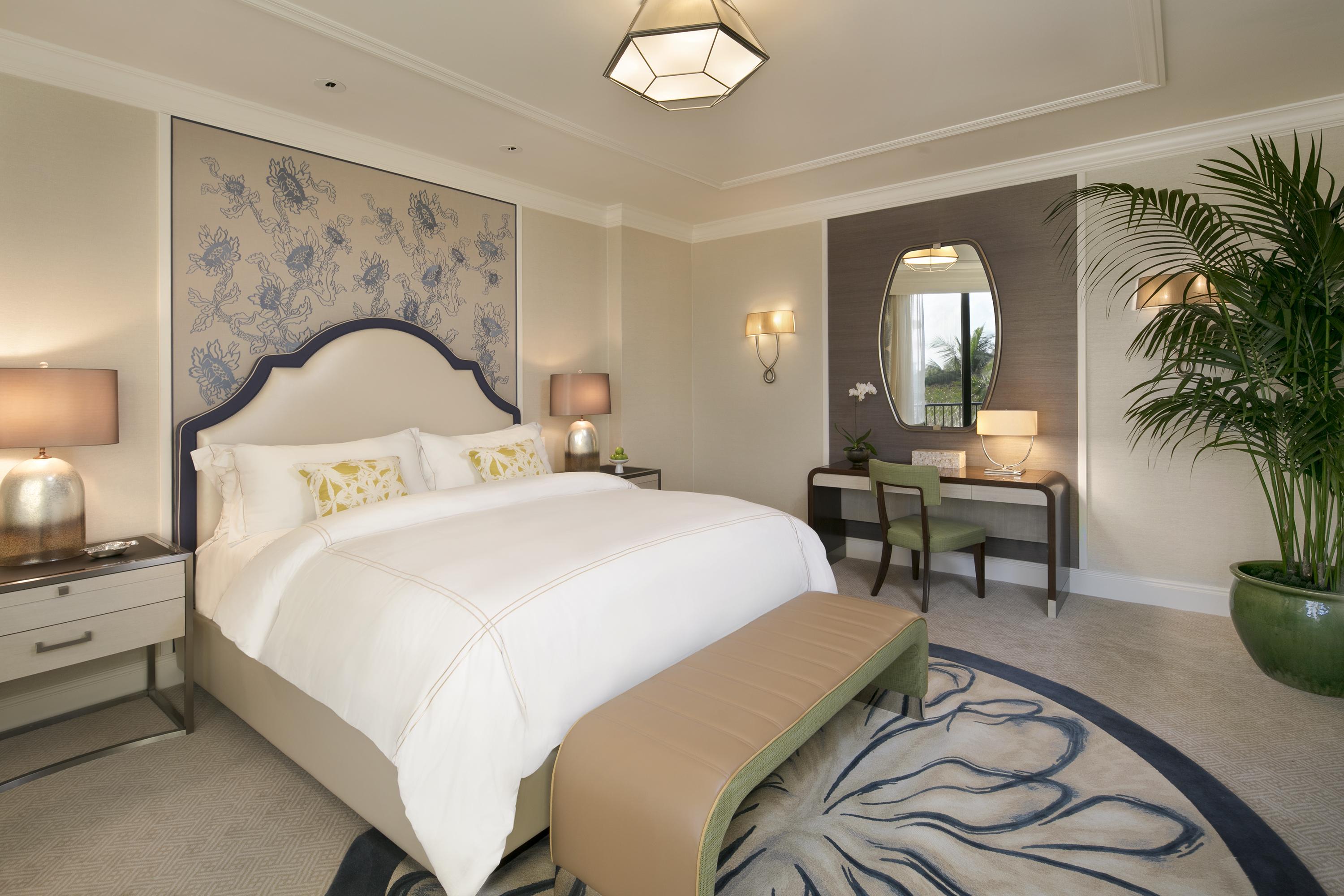 Elegant hotel bedroom with white bedding, blue patterned headboard, vanity desk, and palm plants.