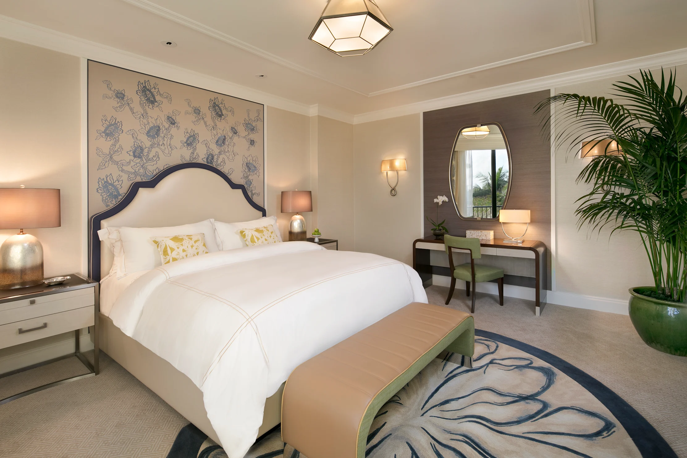 Elegant hotel bedroom with white bedding, blue patterned headboard, vanity desk, and palm plants.