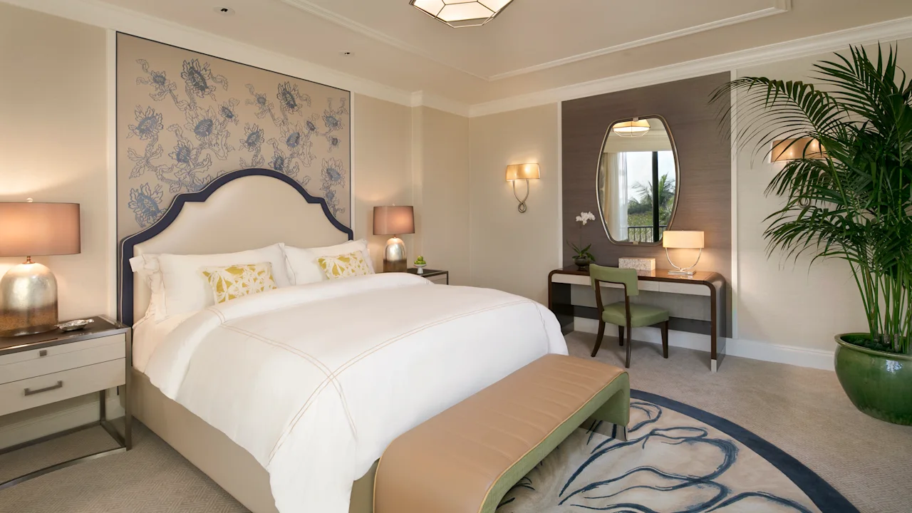 Elegant hotel bedroom with white bedding, blue patterned headboard, vanity desk, and palm plants.