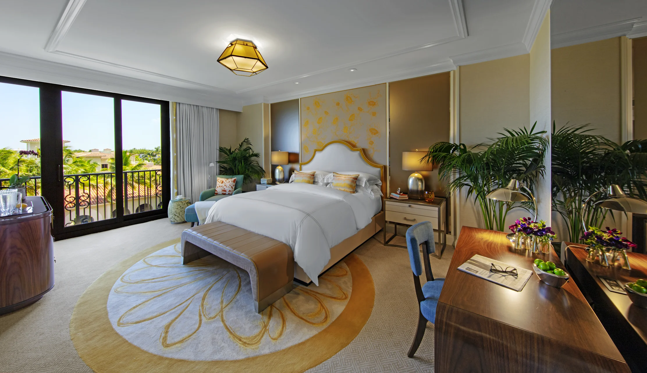 Luxury hotel bedroom with white bedding, gold accents, palm plants, and balcony overlooking greenery.