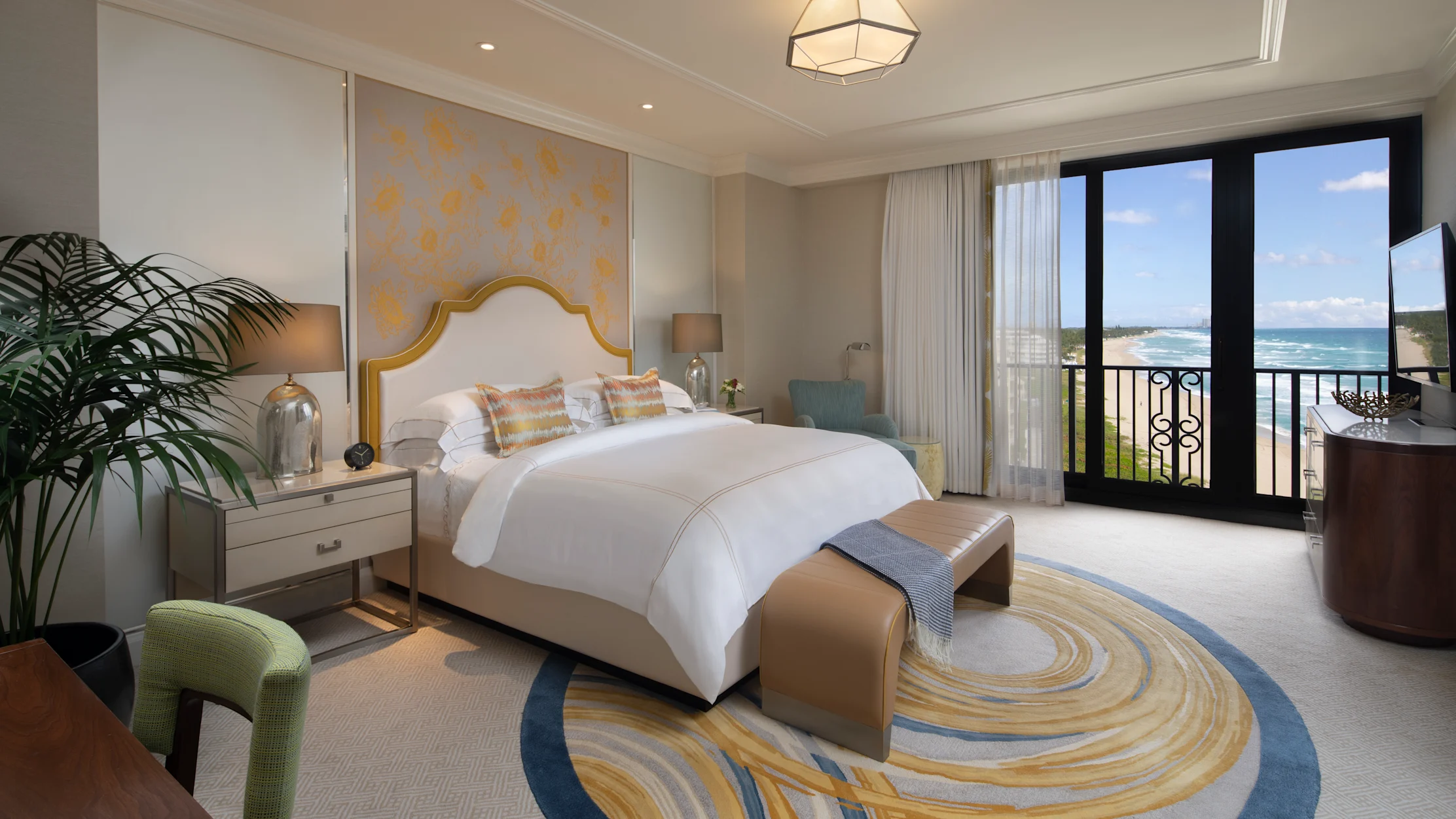 Elegant hotel bedroom with white bed, gold accents, geometric light fixture, and ocean view balcony.