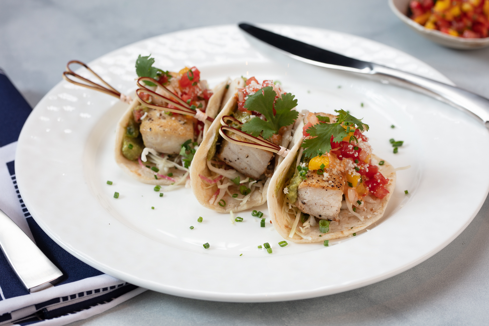 Three fish tacos on a white plate topped with salsa, cilantro, and slaw, garnished with chives.