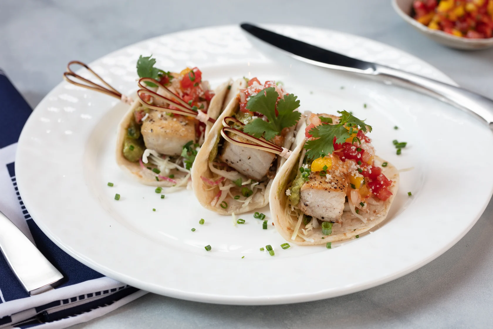 Three fish tacos on a white plate topped with salsa, cilantro, and slaw, garnished with chives.
