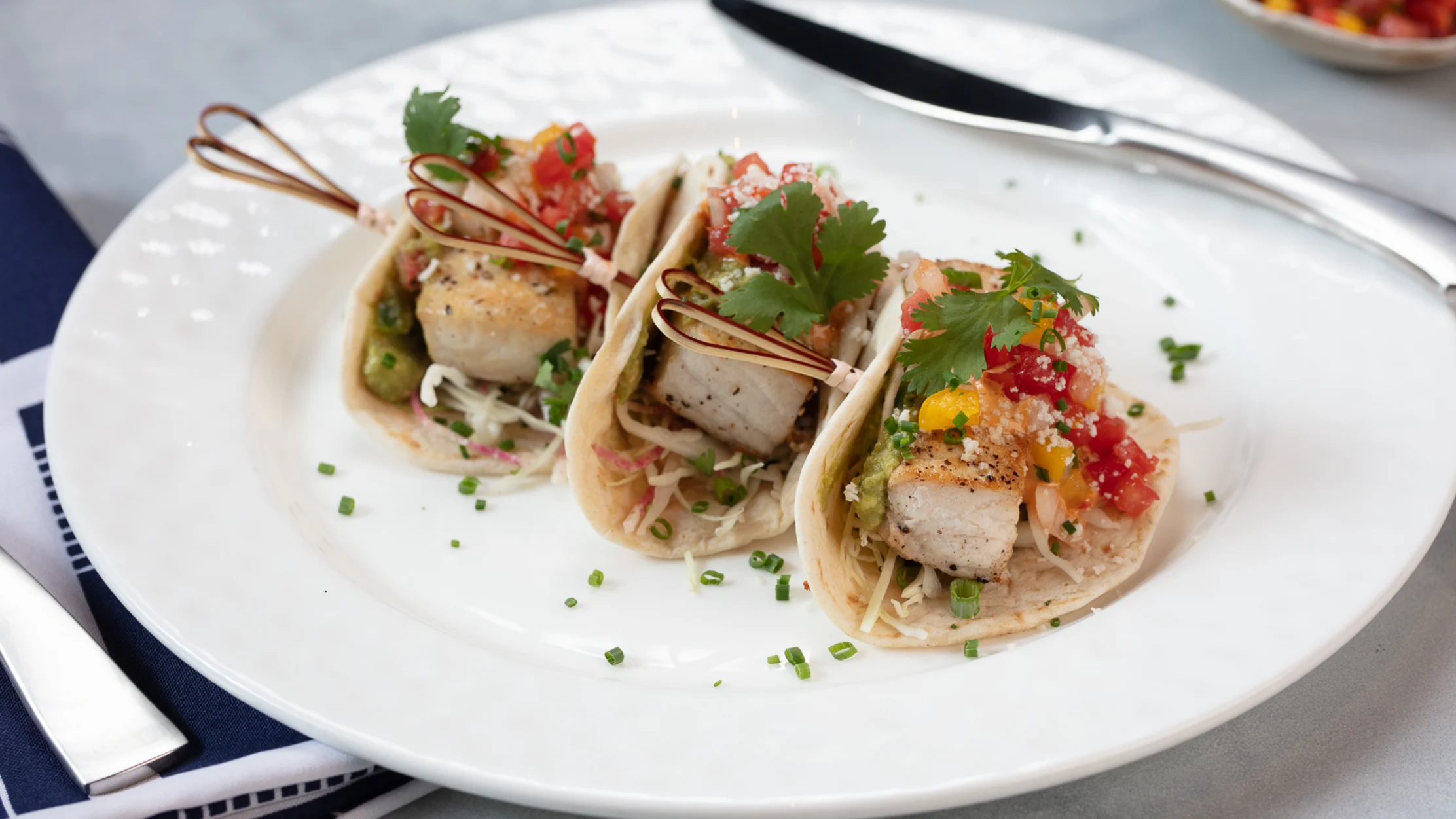 Three fish tacos on a white plate topped with salsa, cilantro, and slaw, garnished with chives.