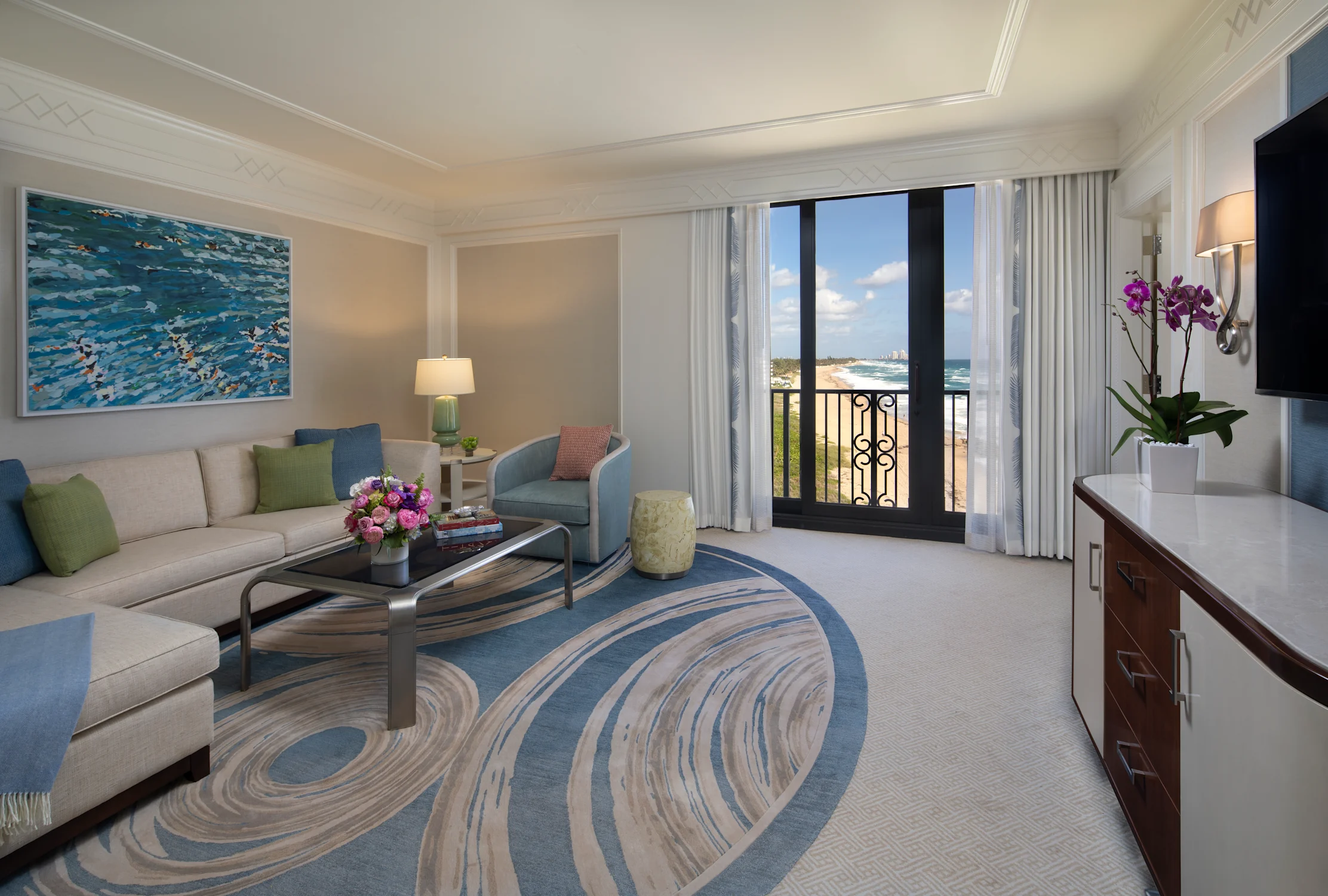Elegant hotel living room with ocean view balcony, blue and beige decor, and coastal artwork.