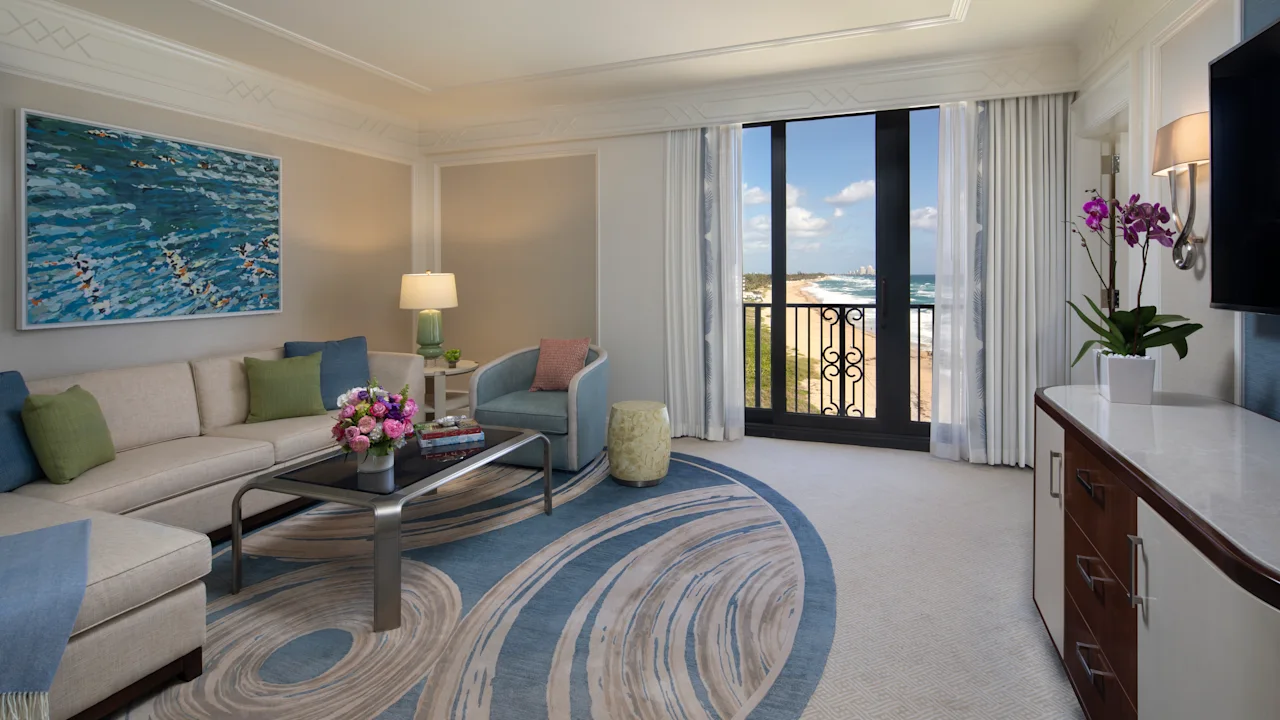 Elegant hotel living room with ocean view balcony, blue and beige decor, and coastal artwork.