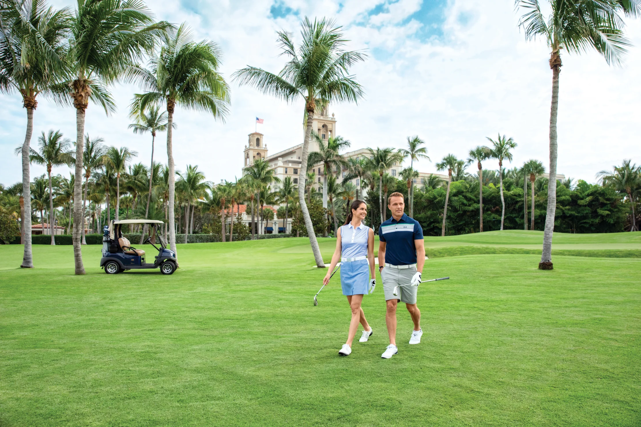 Couple walking on tropical golf course with palm trees, golf cart, and resort building in background.