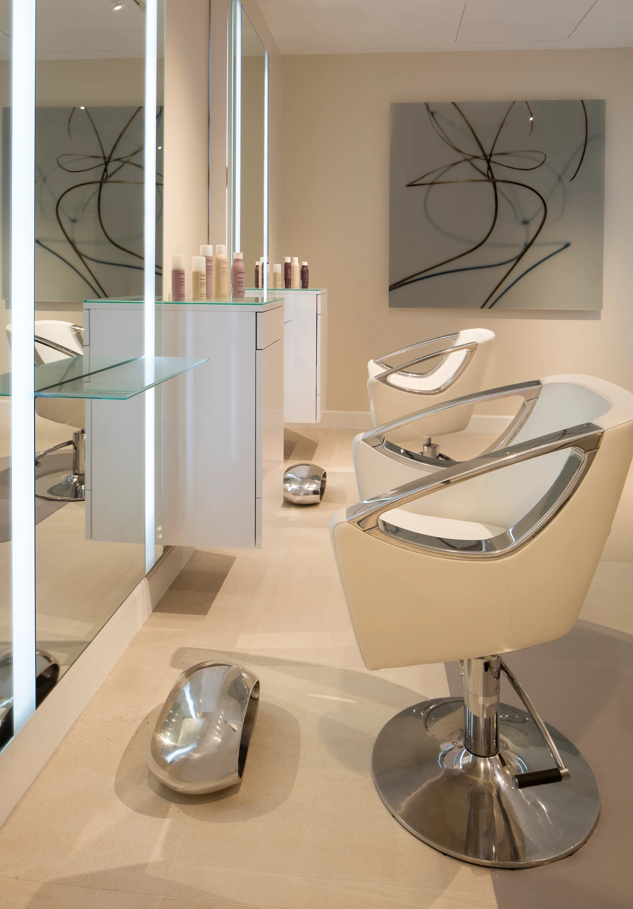 Modern salon interior with beige styling chairs, glass shelving, abstract wall art, and neutral decor.