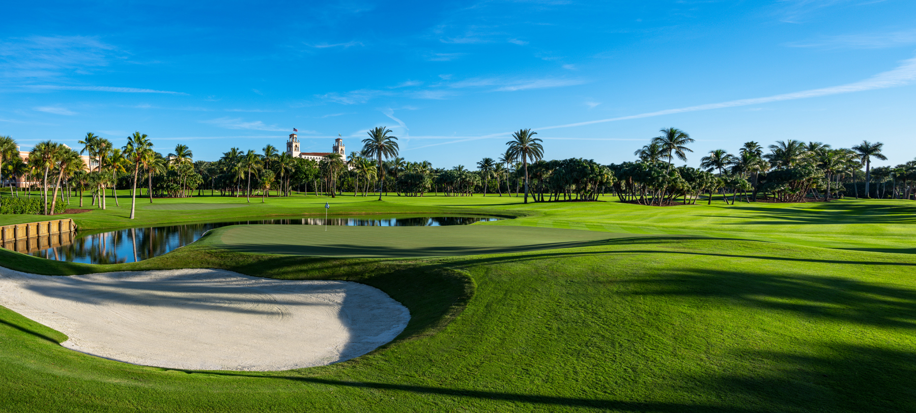 Lush green golf course with sand bunker, water hazard, palm trees, and Spanish-style building under blue sky.