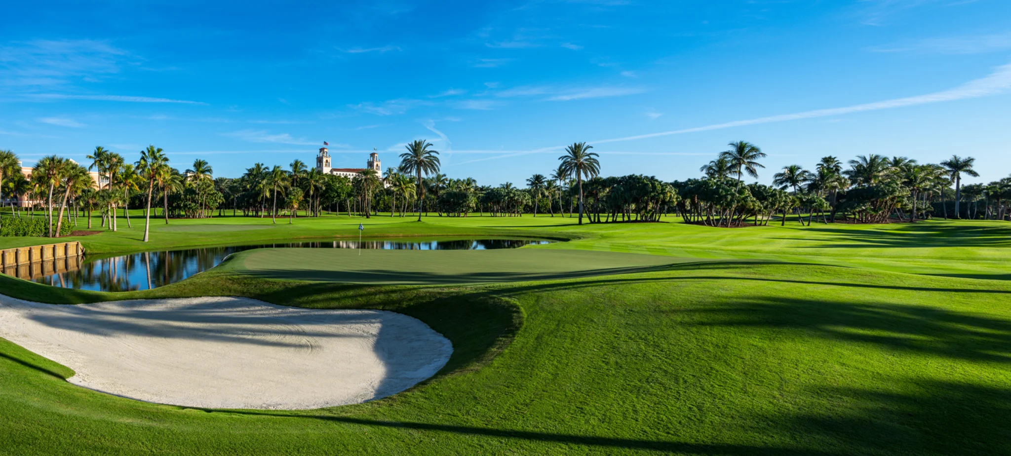 Lush green golf course with sand bunker, water hazard, palm trees, and Spanish-style building under blue sky.