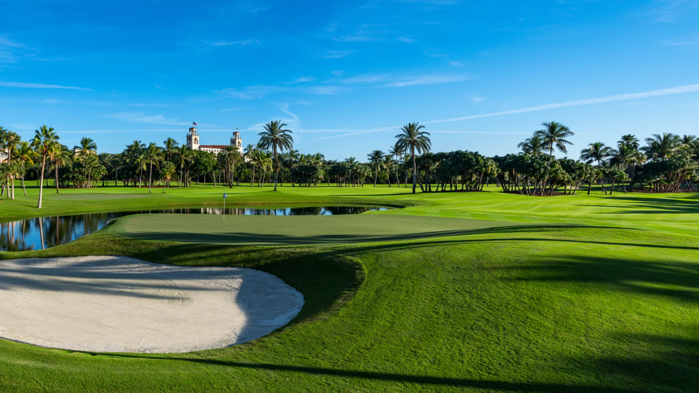 Lush green golf course with sand bunker, water hazard, palm trees, and Spanish-style building under blue sky.