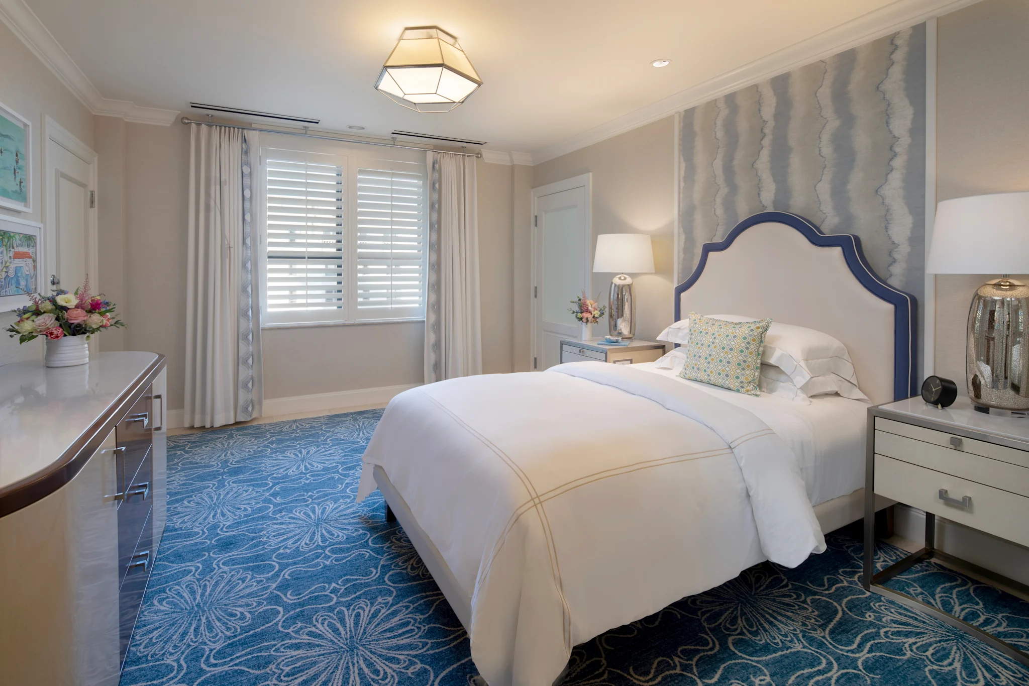 Elegant bedroom with white upholstered bed, blue patterned rug, geometric ceiling light, and neutral decor.