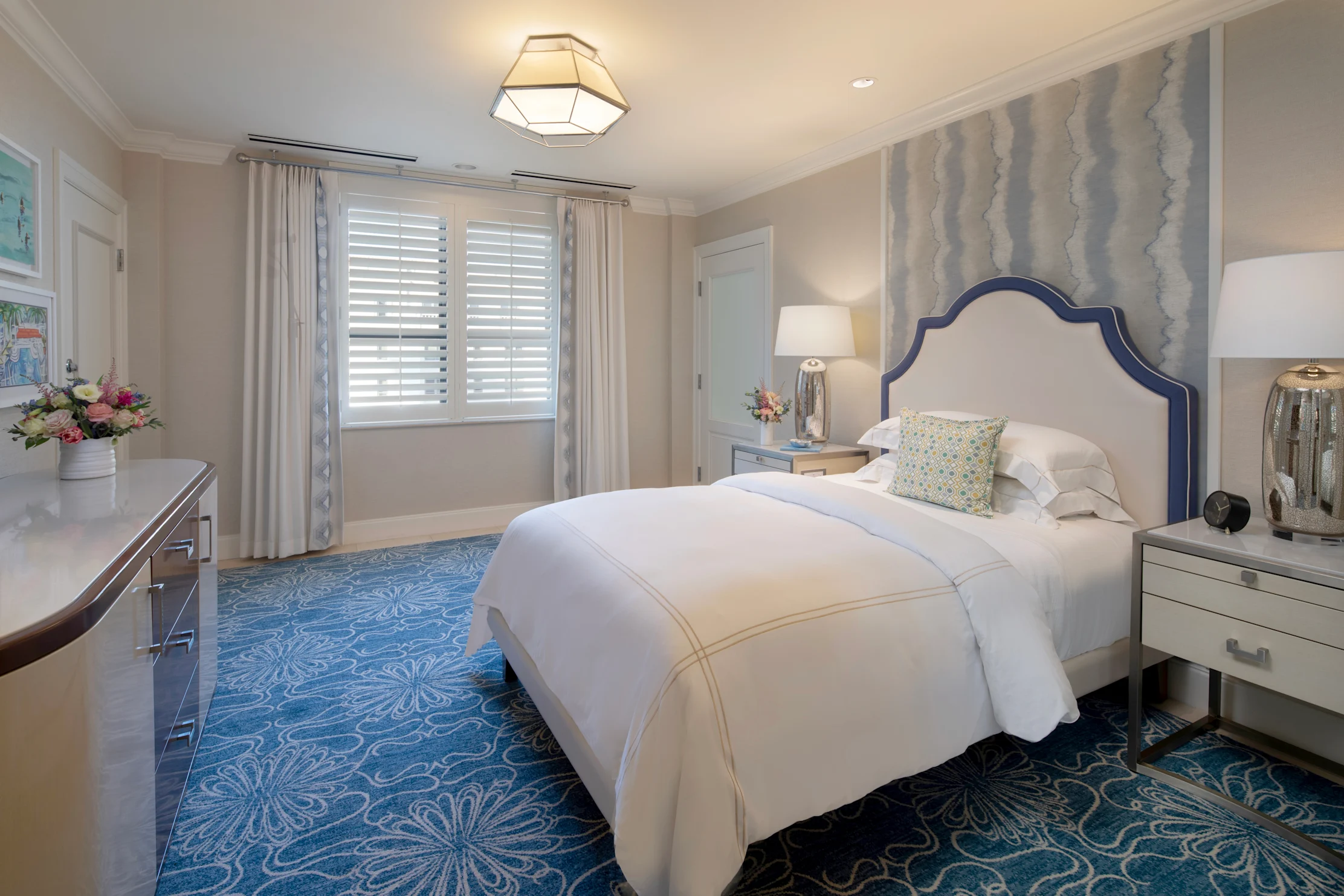 Elegant bedroom with white upholstered bed, blue patterned rug, geometric ceiling light, and neutral decor.
