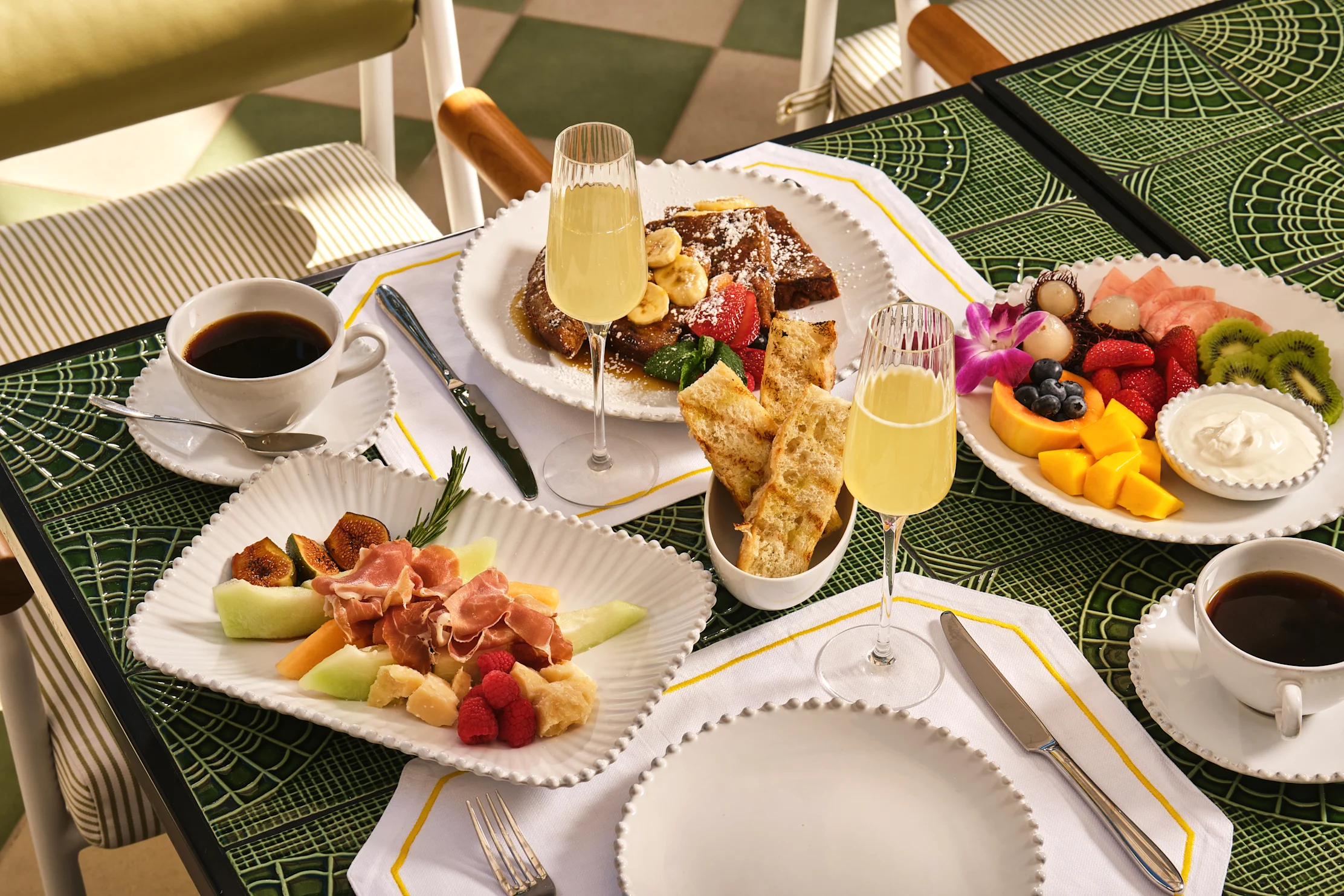 Elegant breakfast spread with French toast, fresh fruit, charcuterie, mimosas and coffee on green table.