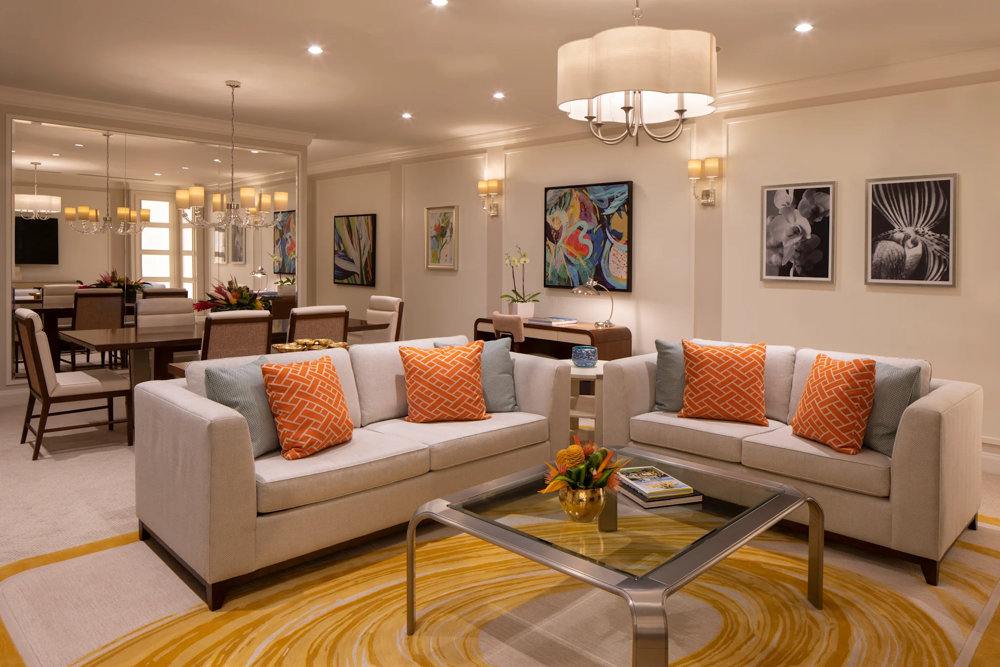 Modern living room with beige sofas, orange pillows, glass coffee table, and colorful artwork.