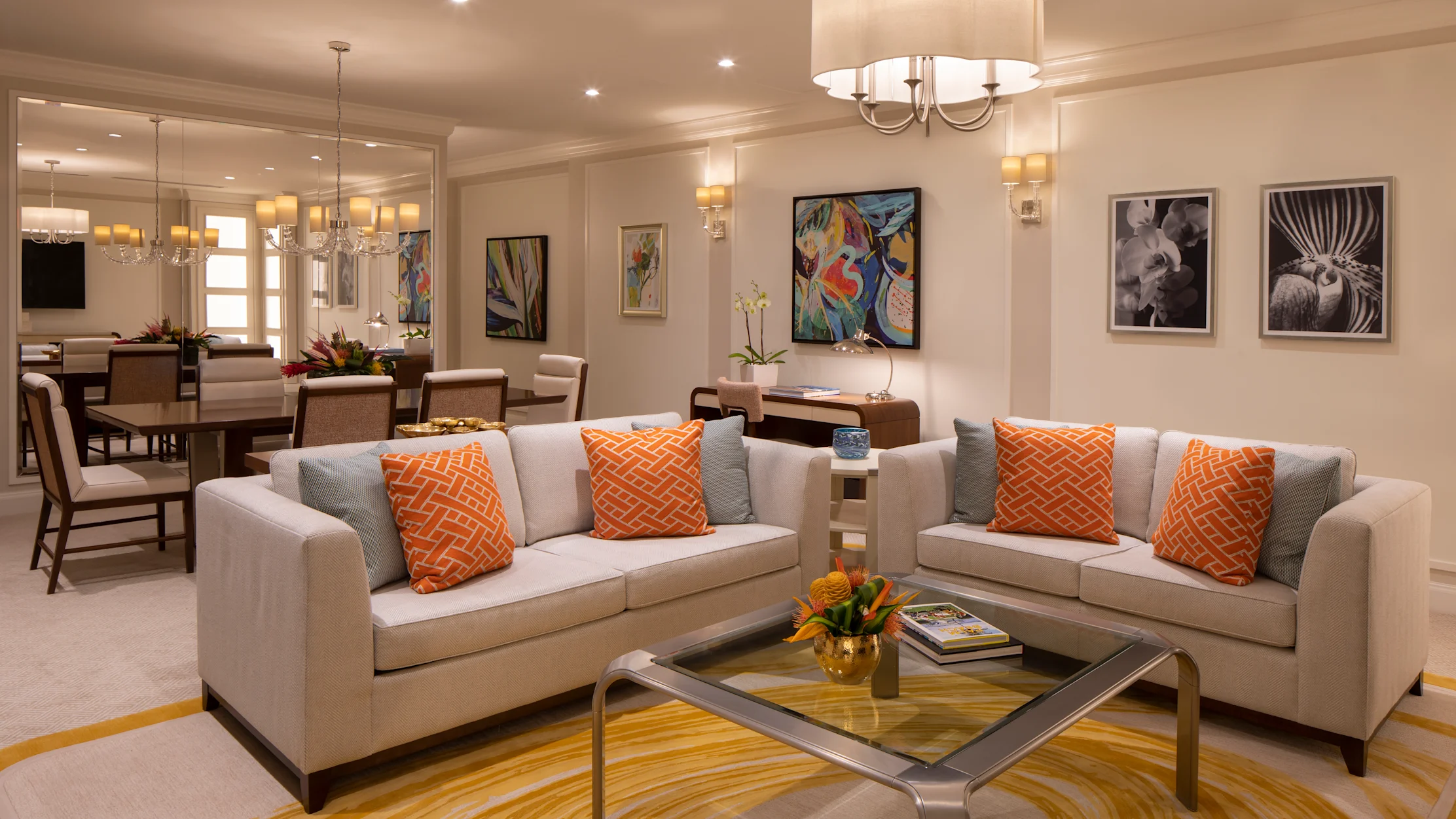 Modern living room with beige sofas, orange pillows, glass coffee table, and colorful artwork.