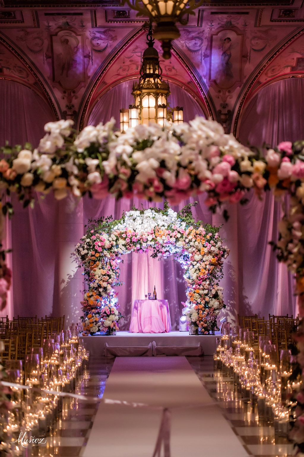 Elegant wedding ceremony venue with floral arch, candlelit aisle, and ornate chandelier under purple lighting.