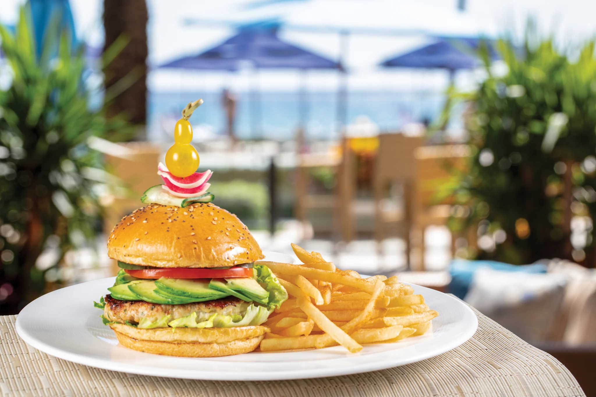 Gourmet burger with avocado and fries on white plate at poolside restaurant with blue umbrellas.