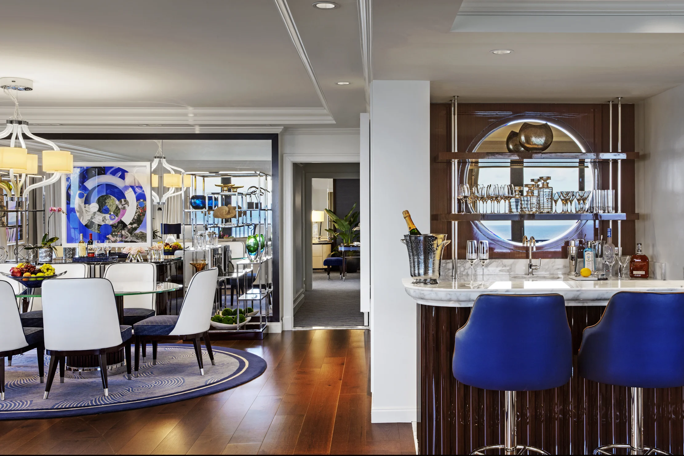Luxury suite interior with dining area, bar with blue stools, and modern artwork visible through doorway.