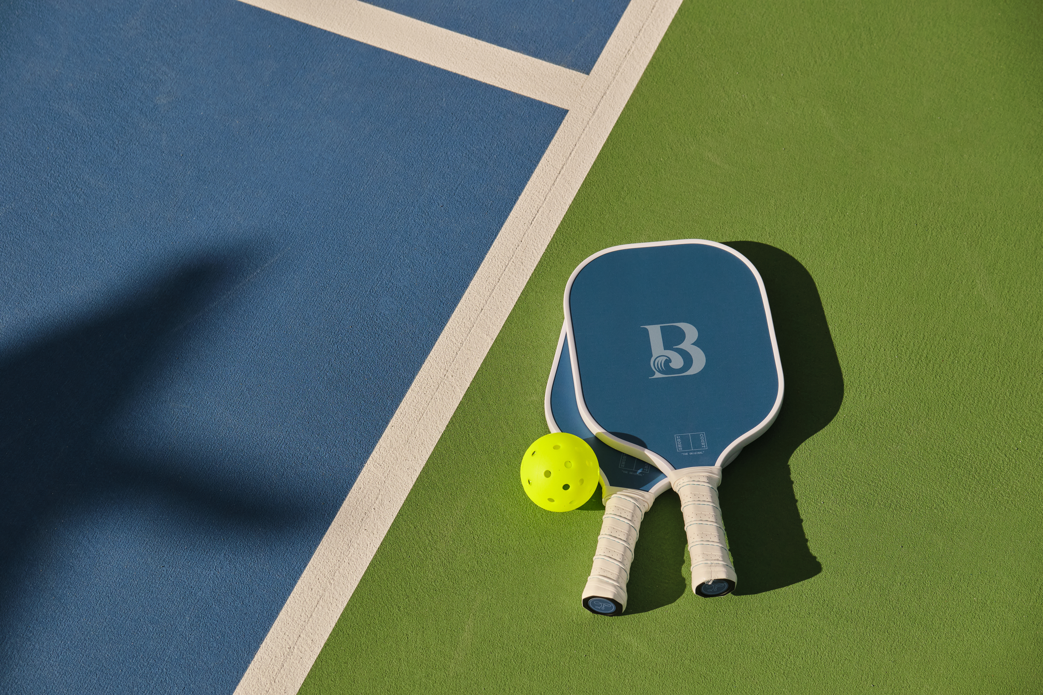 Two blue pickleball paddles and a yellow ball on a green and blue court with white boundary line.
