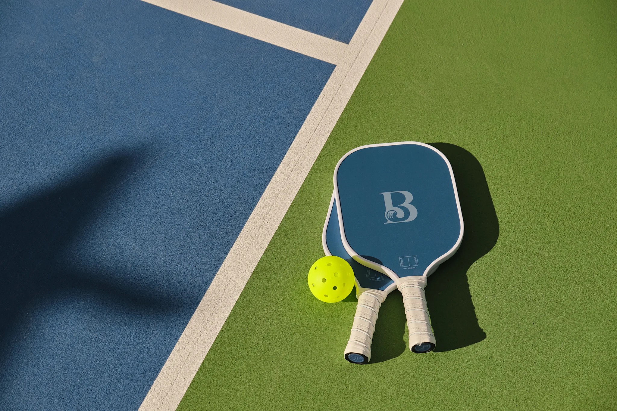 Two blue pickleball paddles and a yellow ball on a green and blue court with white boundary line.