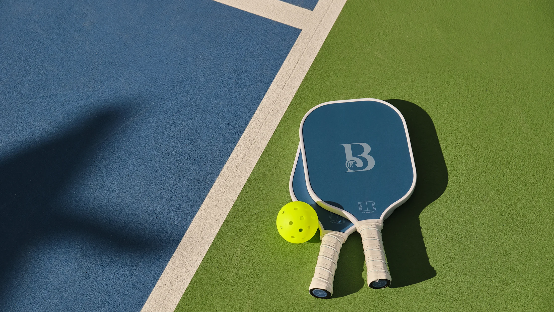 Two blue pickleball paddles and a yellow ball on a green and blue court with white boundary line.