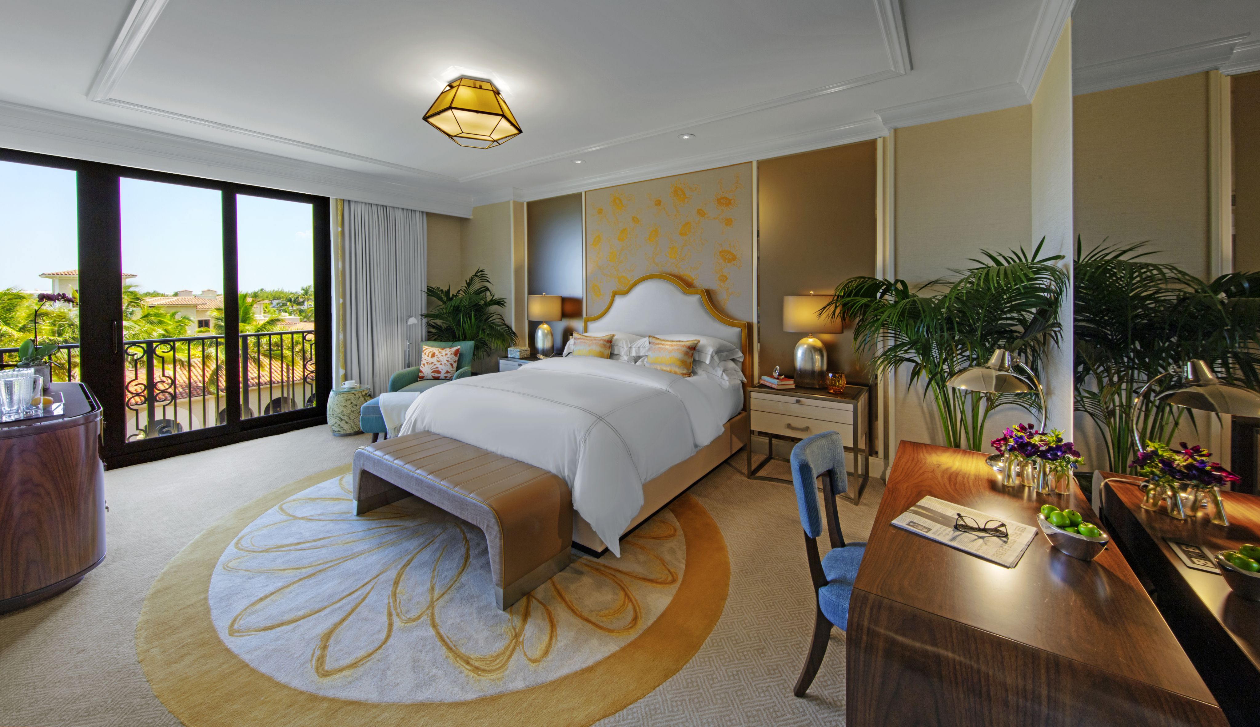 Luxury hotel bedroom with white bedding, gold accents, palm plants, and balcony overlooking resort.