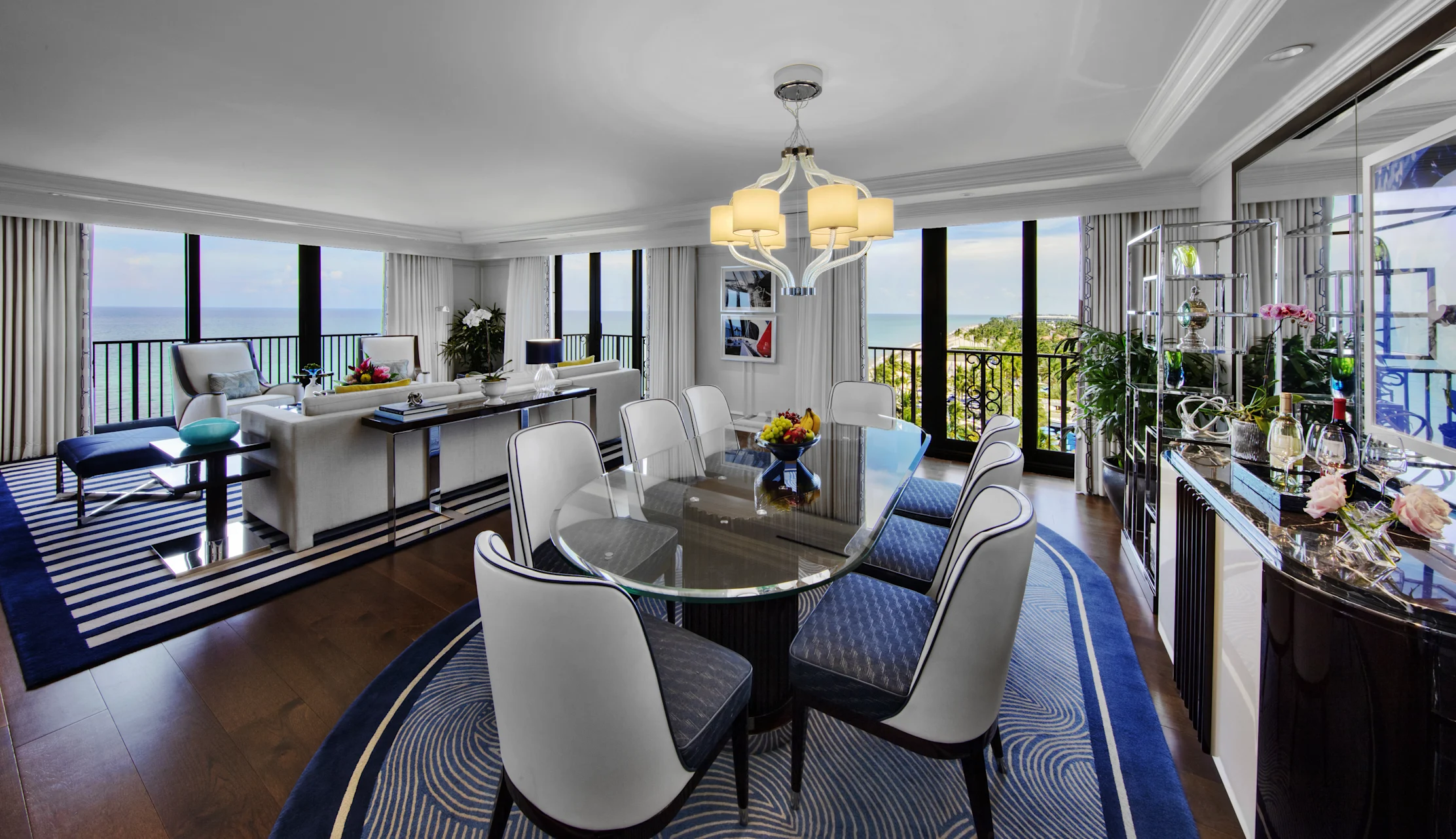 Luxury hotel suite with ocean view, featuring dining area with glass table and living room.