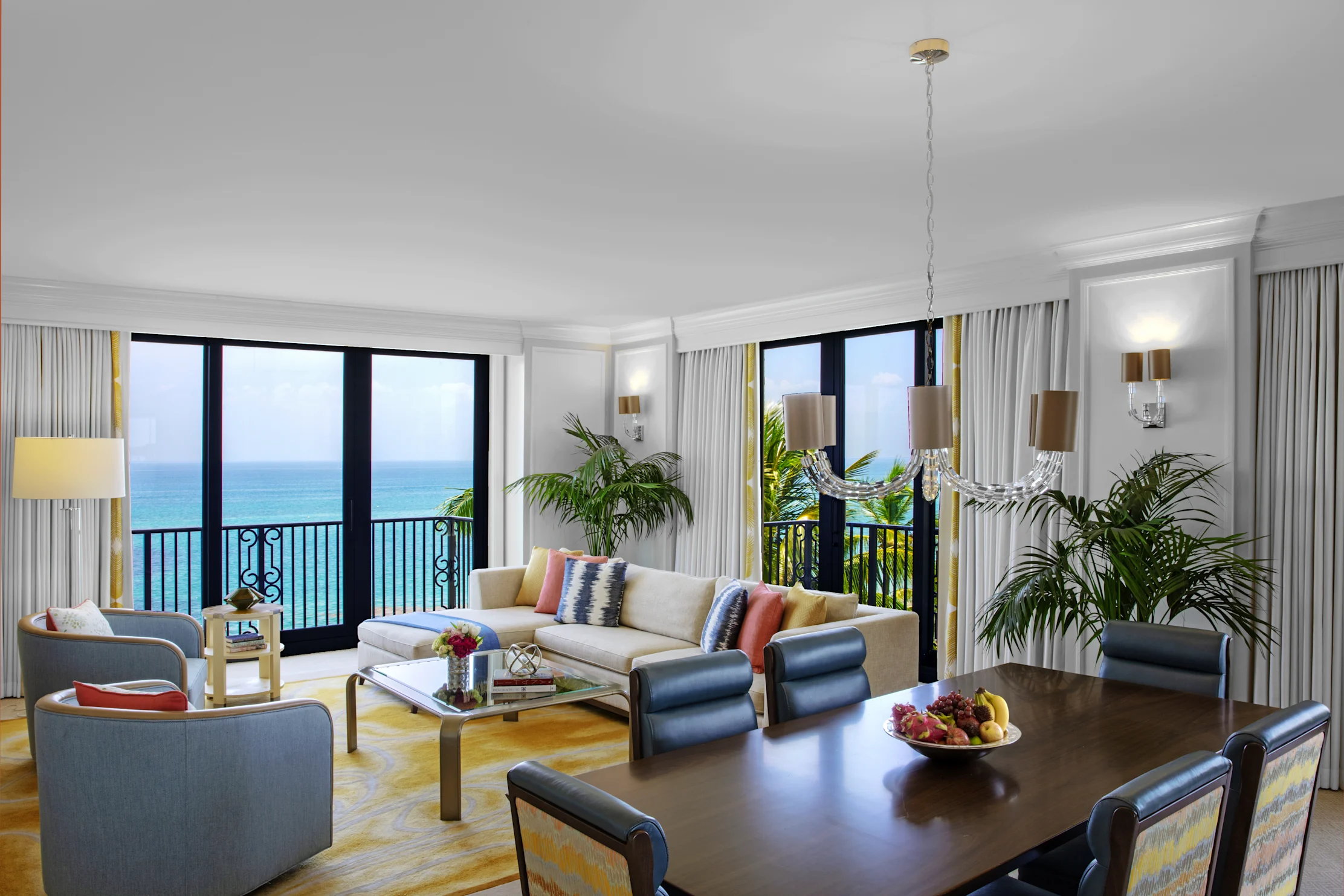 Bright hotel suite with ocean view, white sofa, dining table, and tropical plants by large windows.