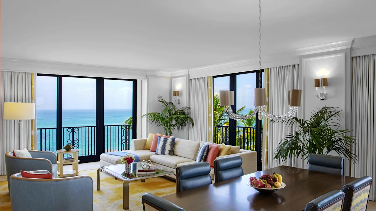 Bright hotel suite with ocean view, white sofa, dining table, and tropical plants by large windows.