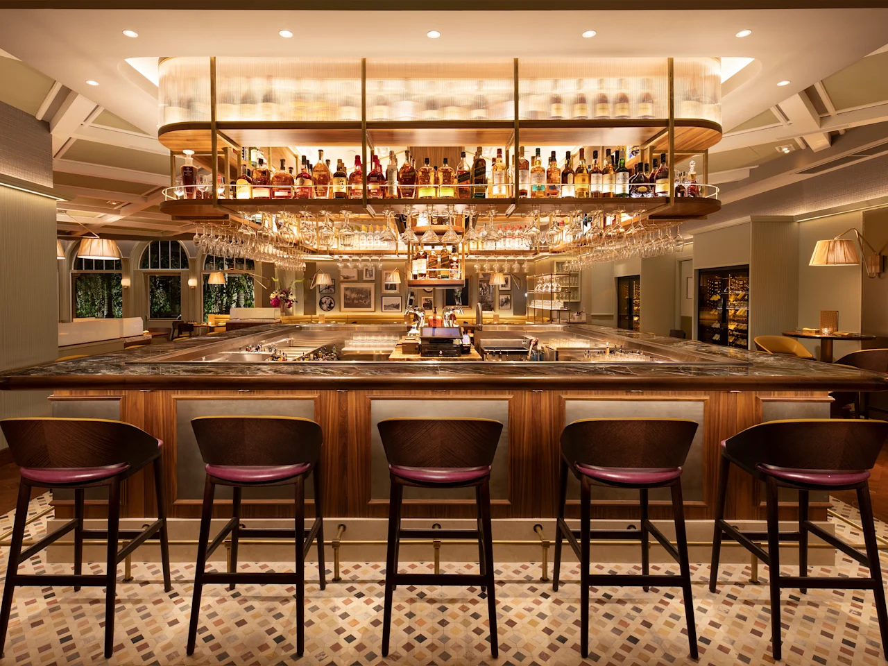 Upscale hotel bar with wooden counter, burgundy leather stools, and illuminated liquor display.