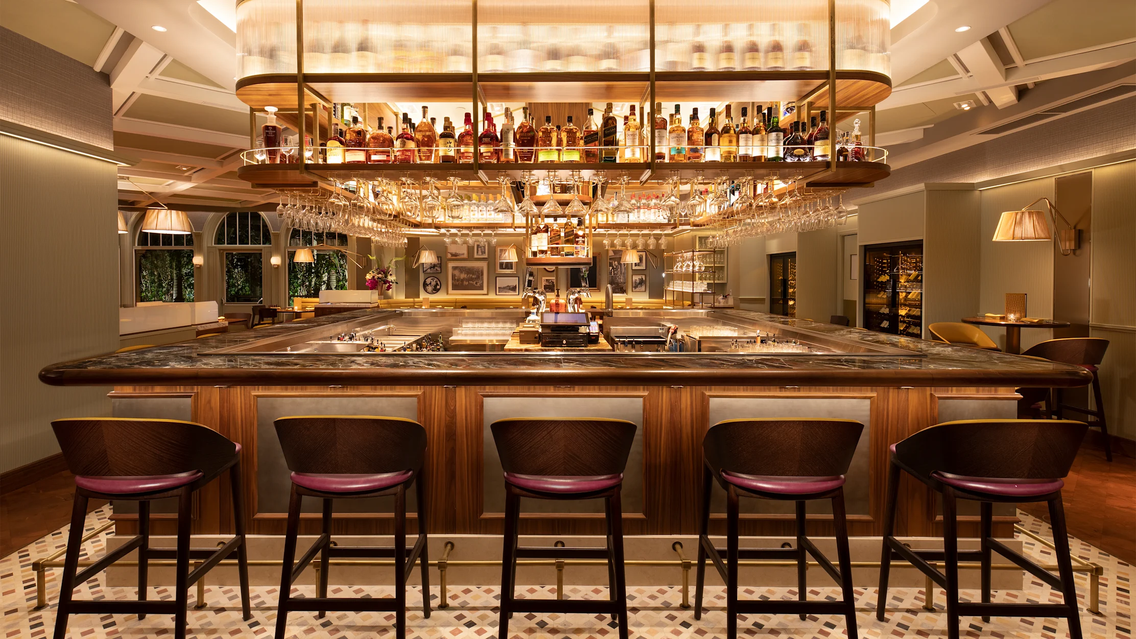 Upscale hotel bar with wooden counter, burgundy leather stools, and illuminated liquor display.