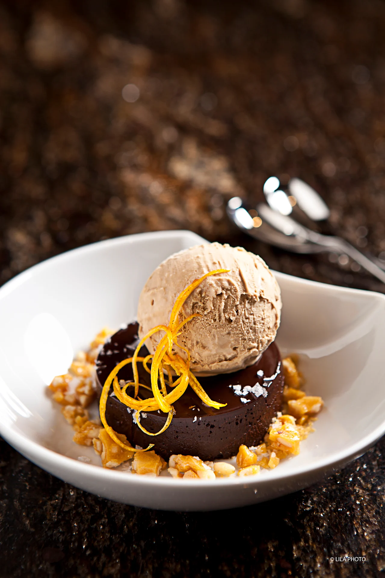 Chocolate brownie topped with ice cream scoop and orange zest on white plate with nuts.