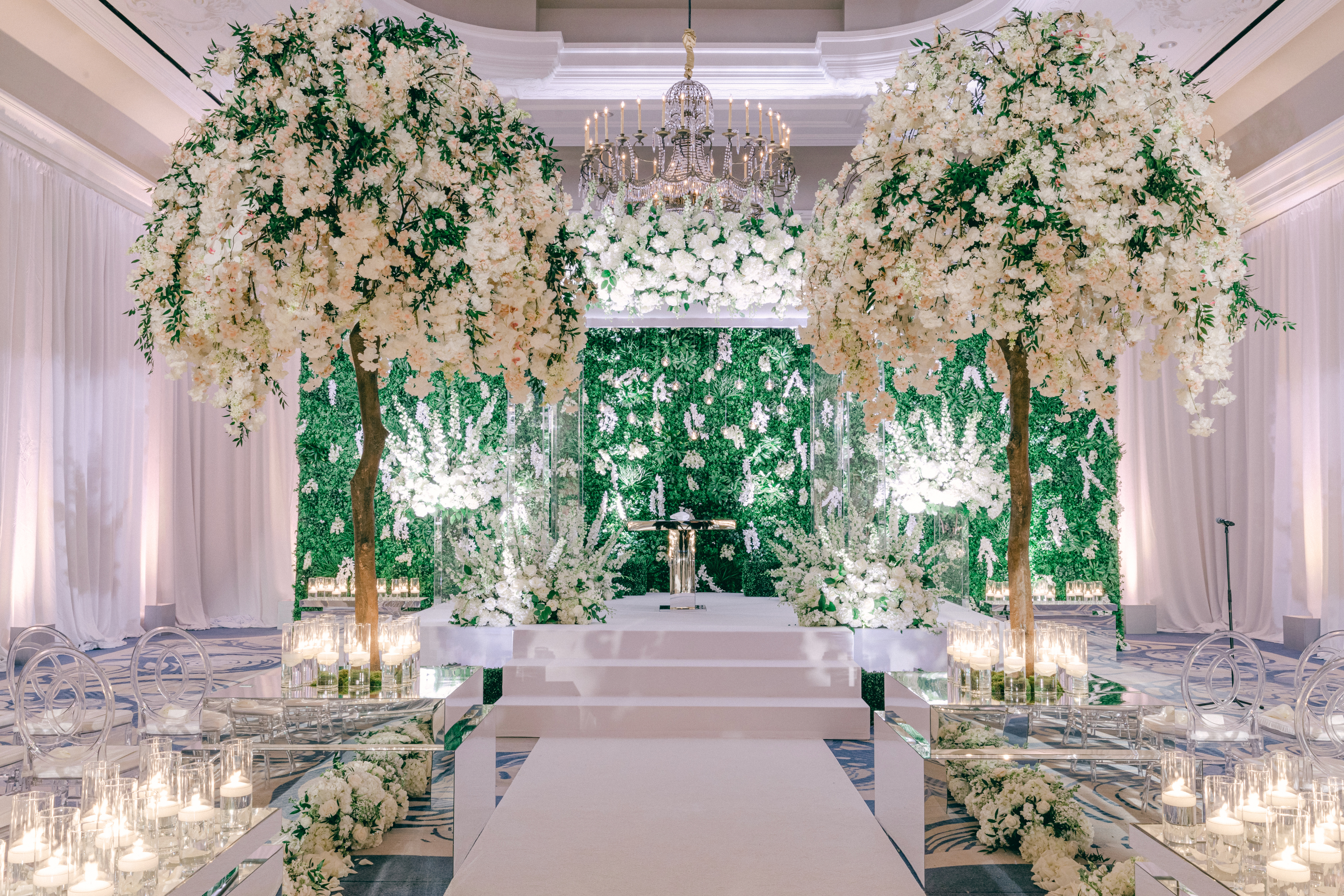 Elegant wedding ceremony setup with white floral trees, green backdrop, chandelier, and candle-lined aisle.