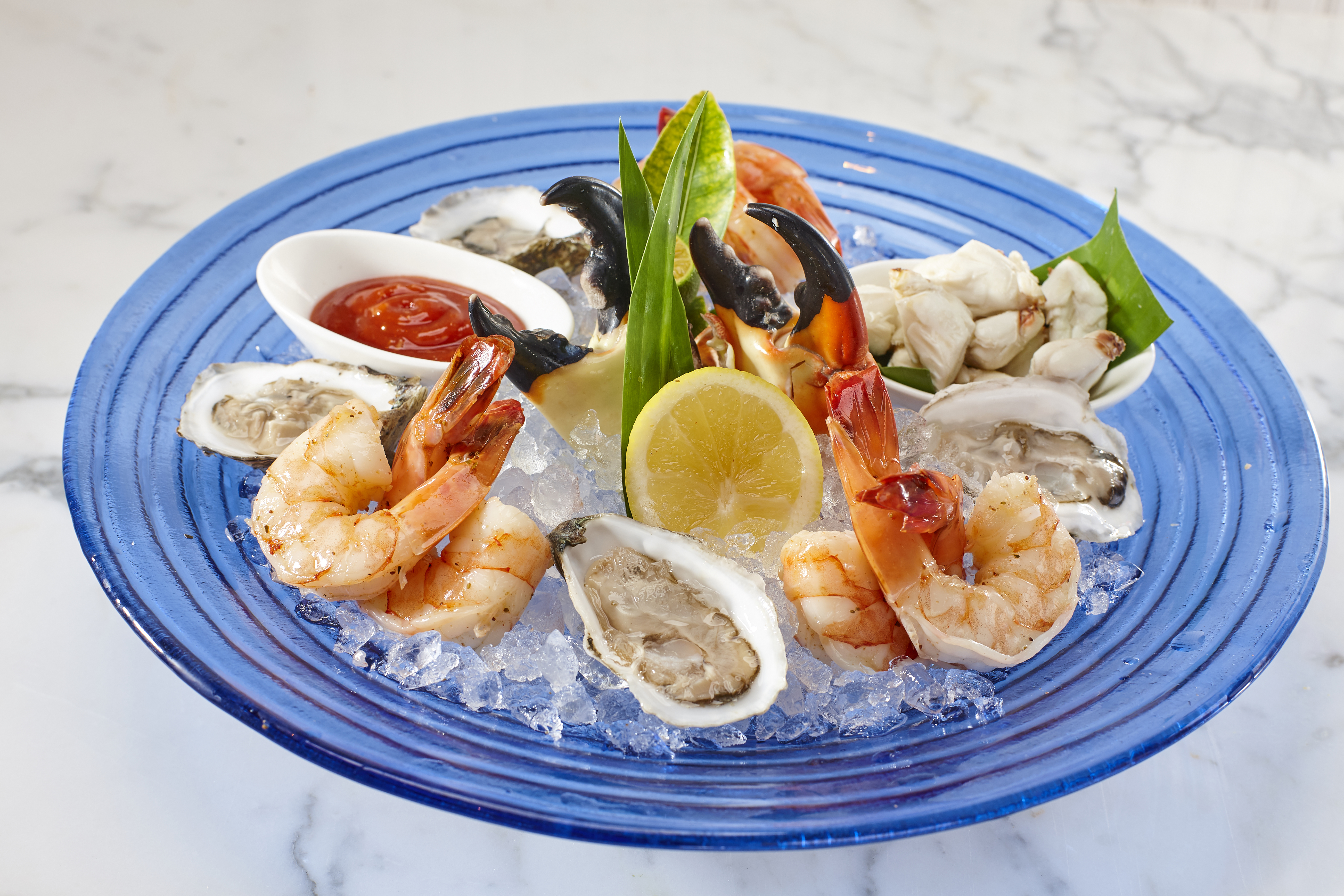 Seafood platter on blue plate with shrimp, oysters, crab, lemon, and cocktail sauce on ice.
