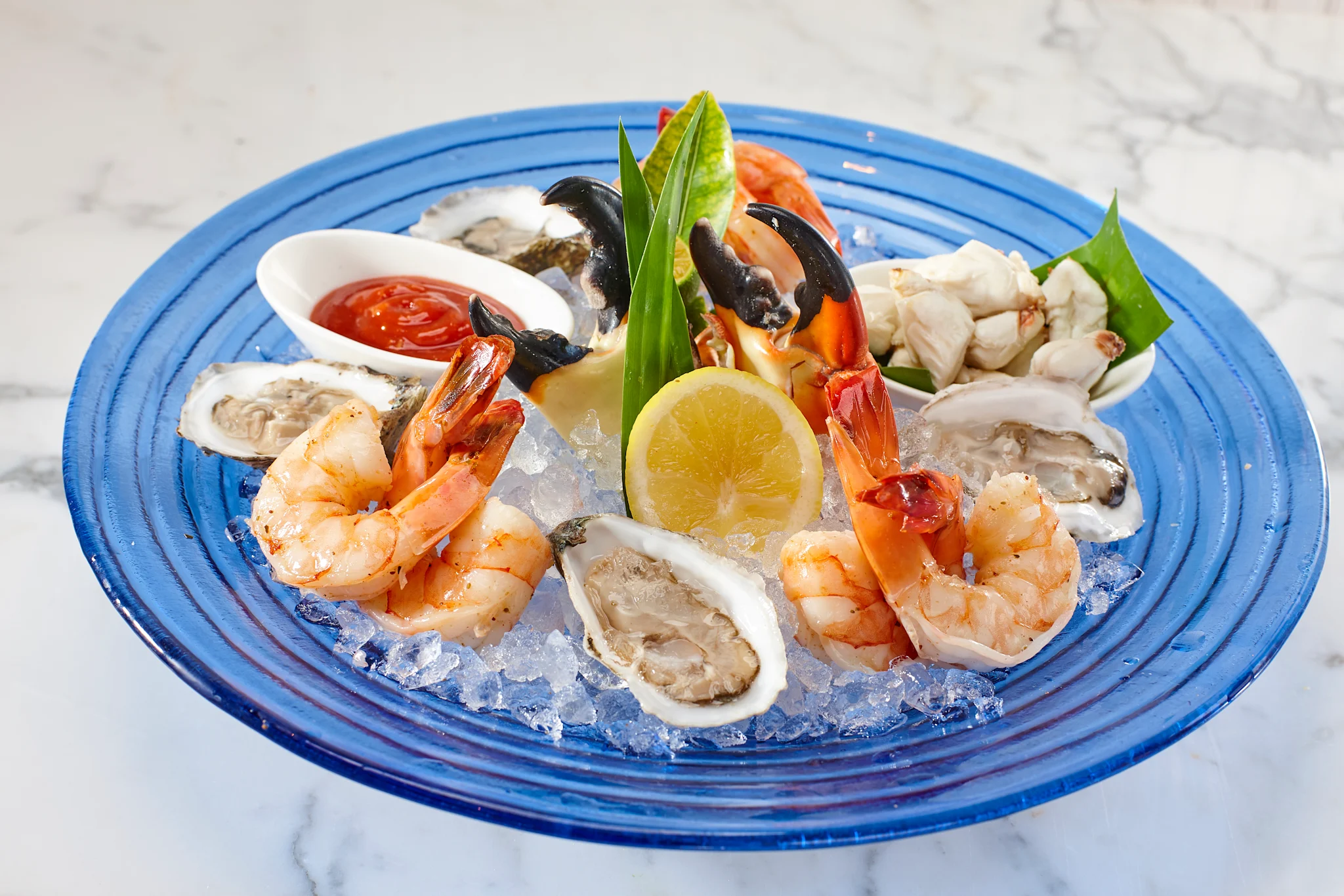 Seafood platter on blue plate with shrimp, oysters, crab, lemon, and cocktail sauce on ice.
