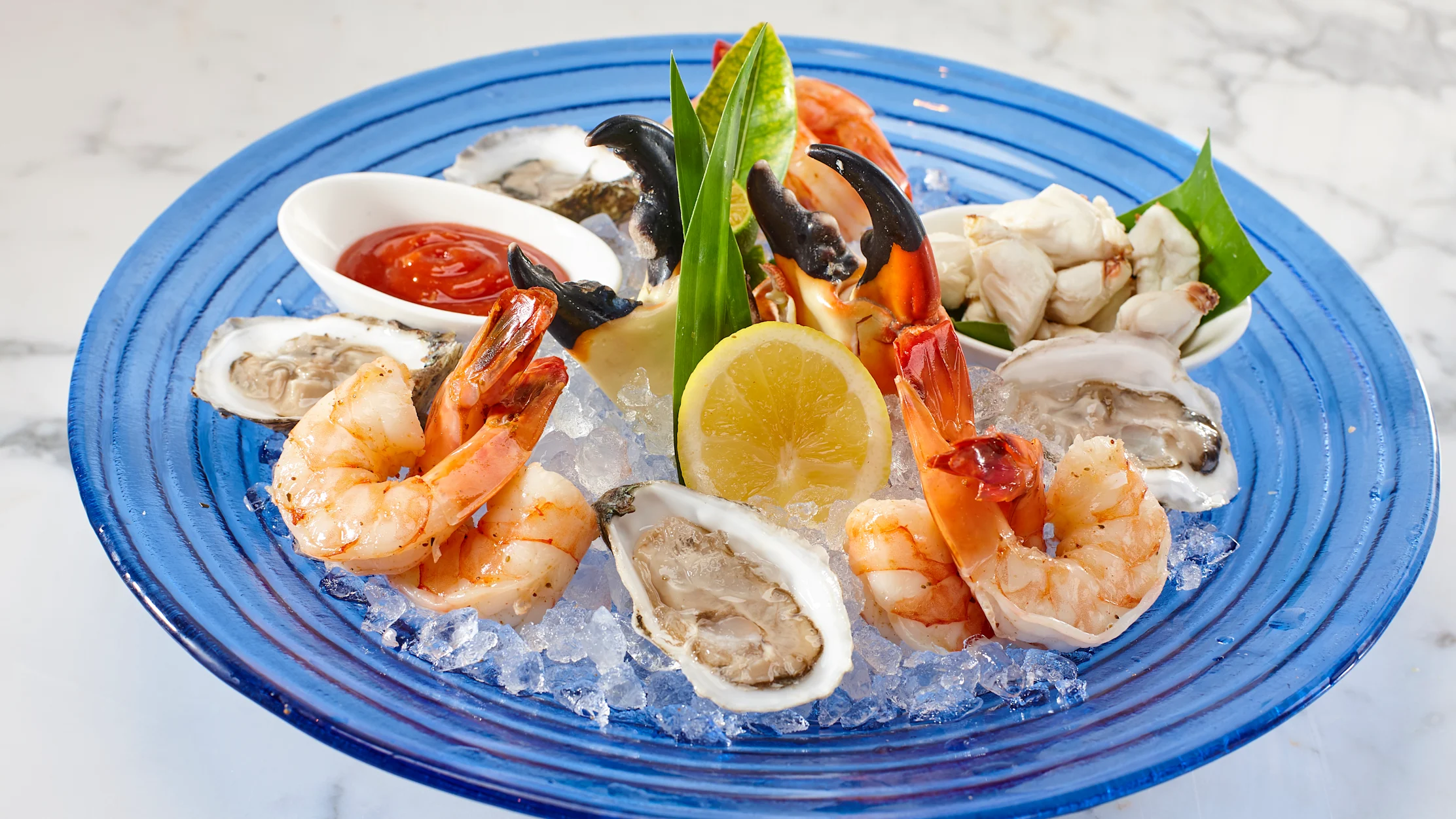 Seafood platter on blue plate with shrimp, oysters, crab, lemon, and cocktail sauce on ice.